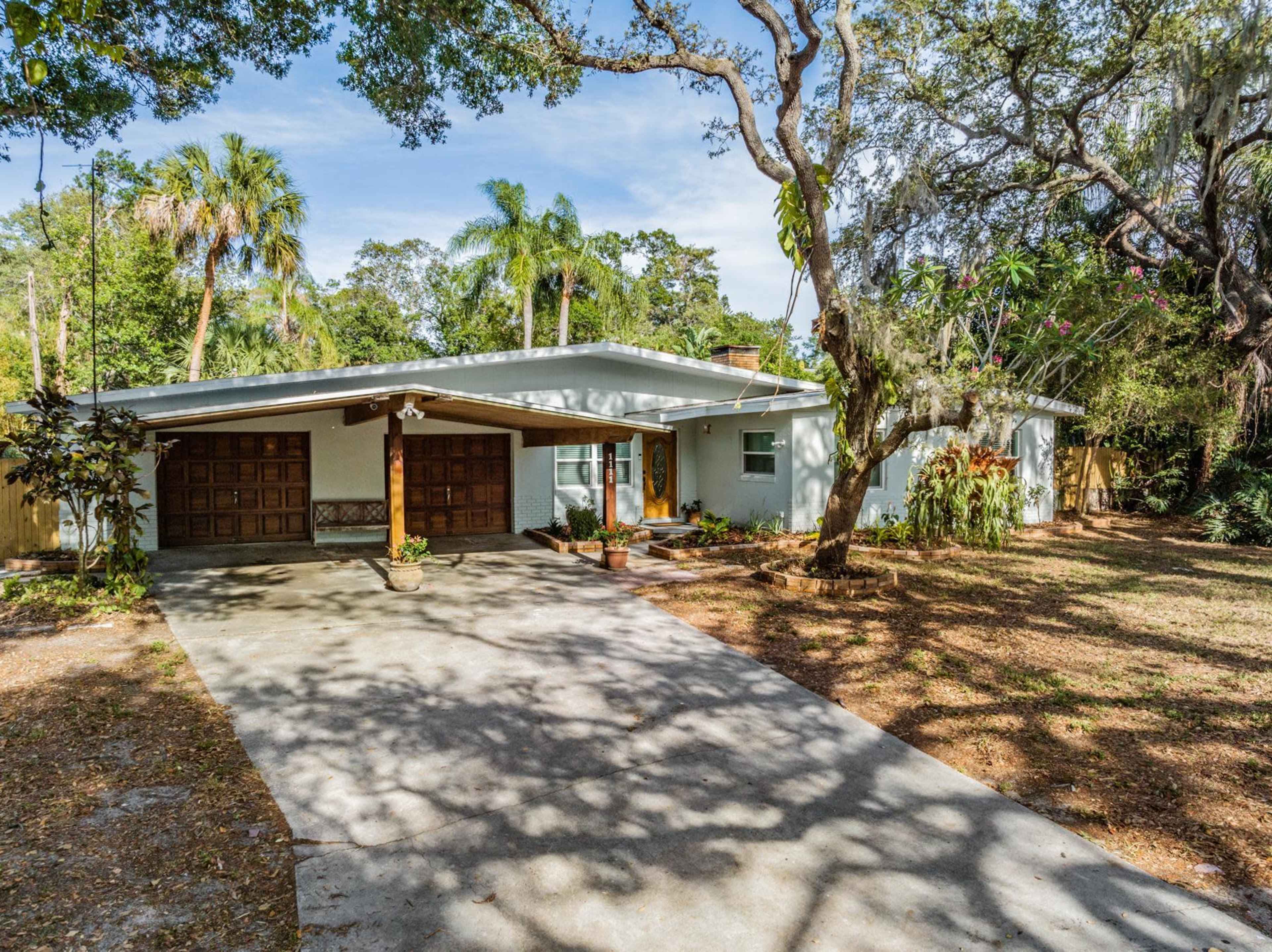 A single-story home with a two-car garage, set in a landscaped yard featuring trees and tropical plants.