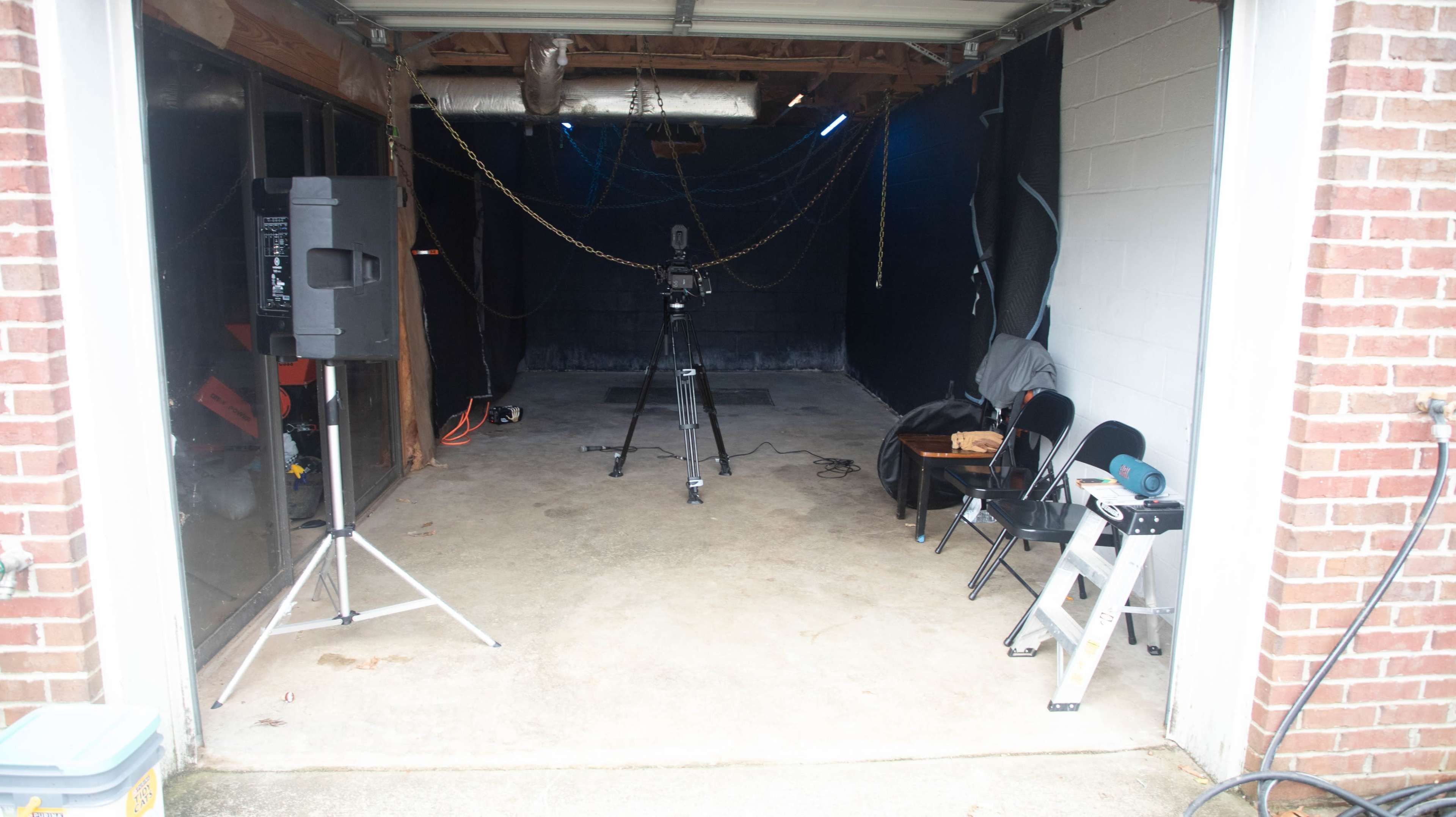 The image shows an empty garage space with a camera on a tripod, black backdrop, and audio equipment set up, alongside a few chairs and a ladder.