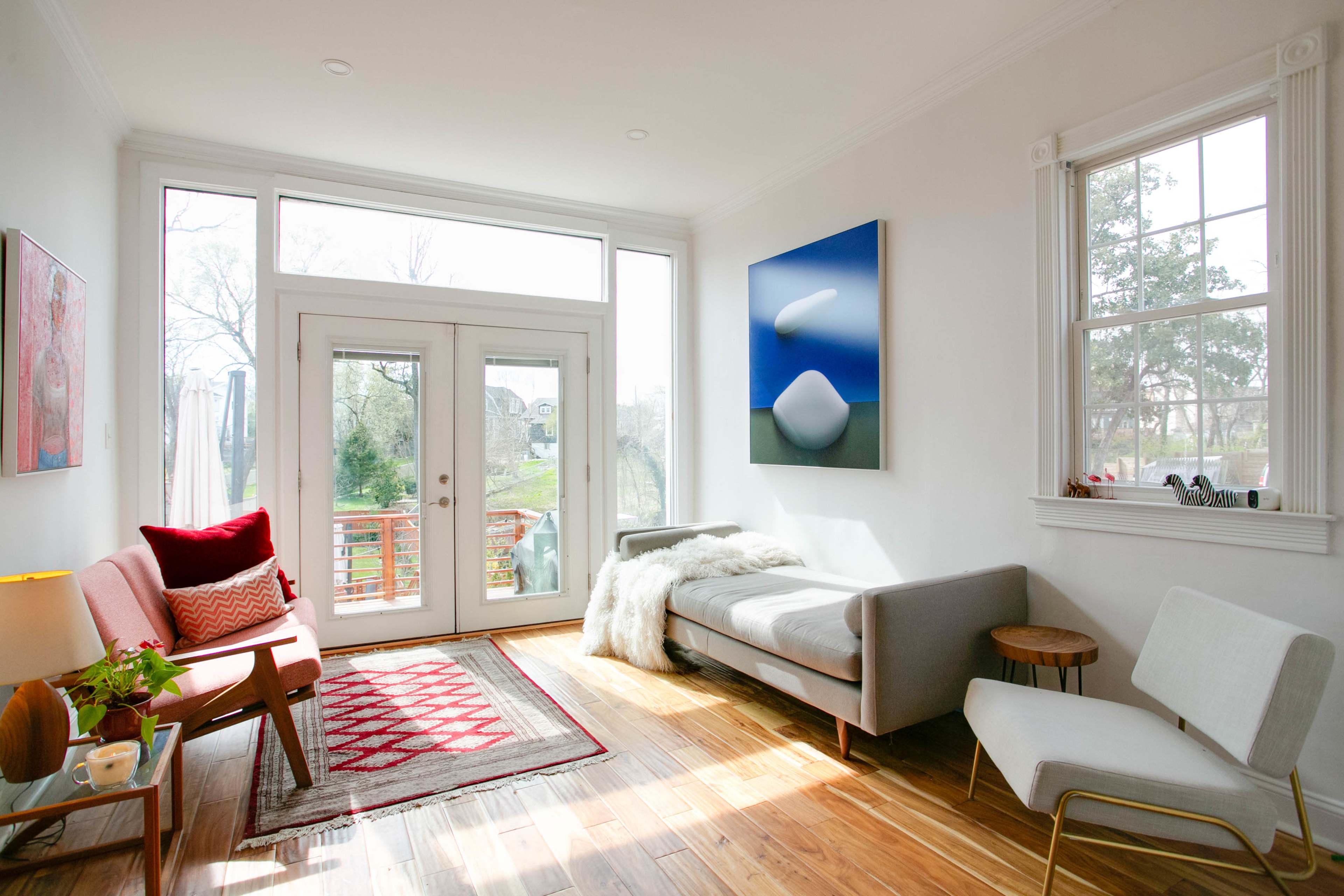 A bright living room features a gray sofa, a red patterned rug, and large windows opening to a view of the outdoors.