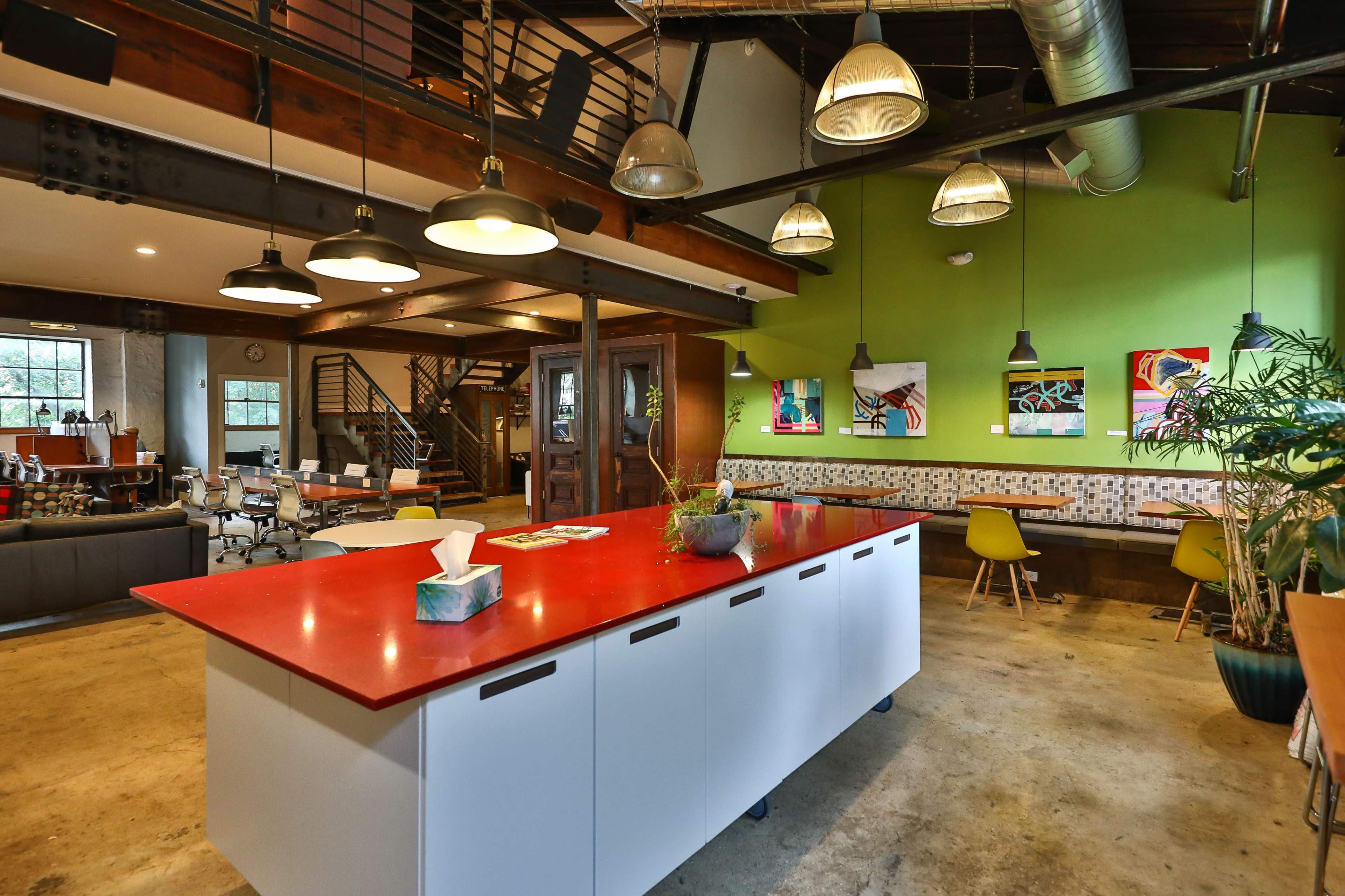 A modern open-concept office space featuring a red countertop island, green accent walls, and various seating areas with artistic decorations.