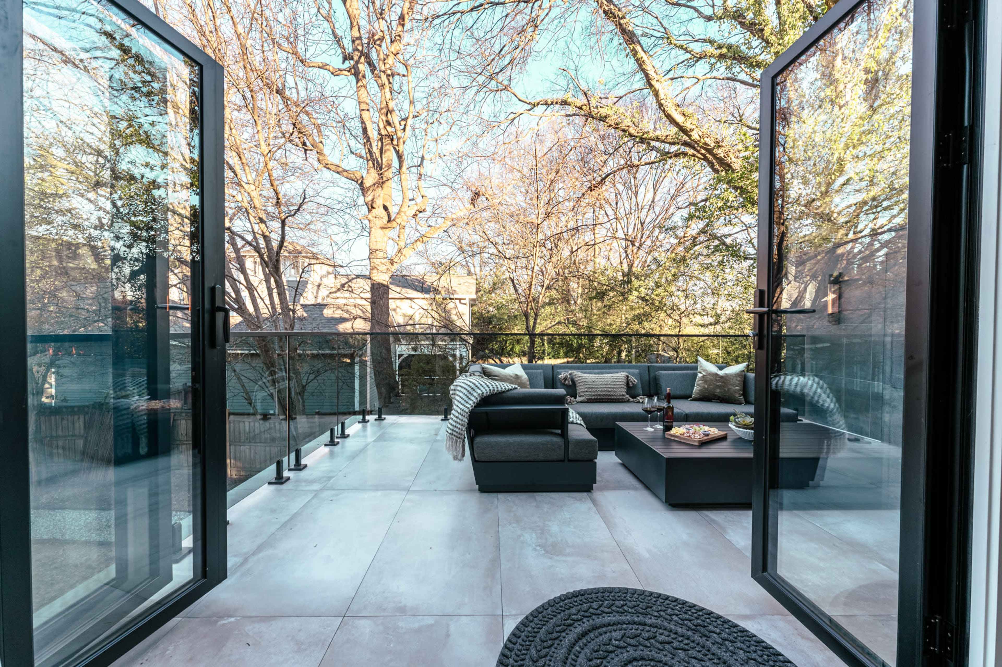 The image shows a spacious balcony with black furniture, a coffee table, and a view of trees and neighboring houses in the background.