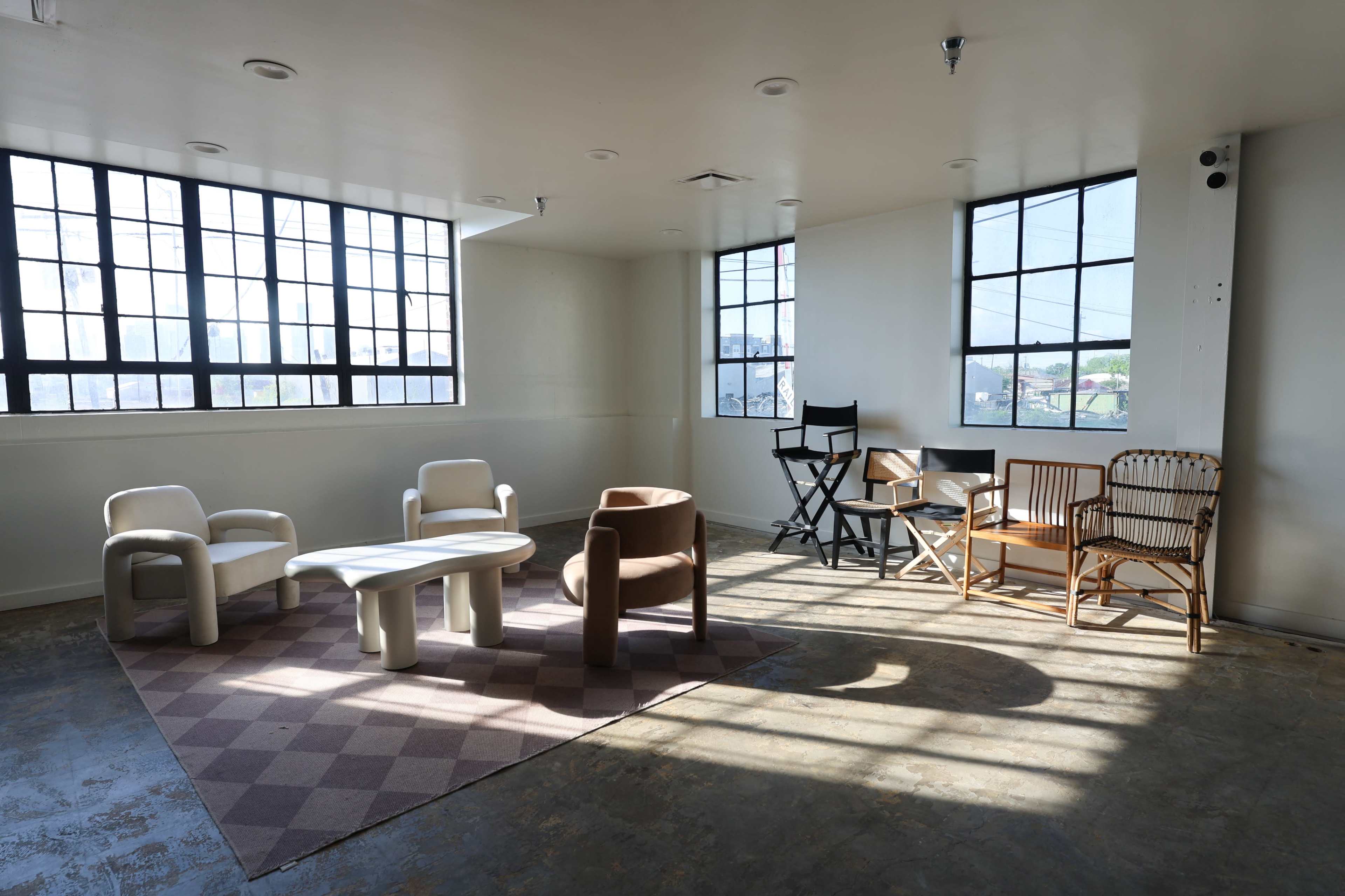 The image shows a spacious, well-lit room with a variety of chairs and a coffee table arranged around a patterned rug near large windows.