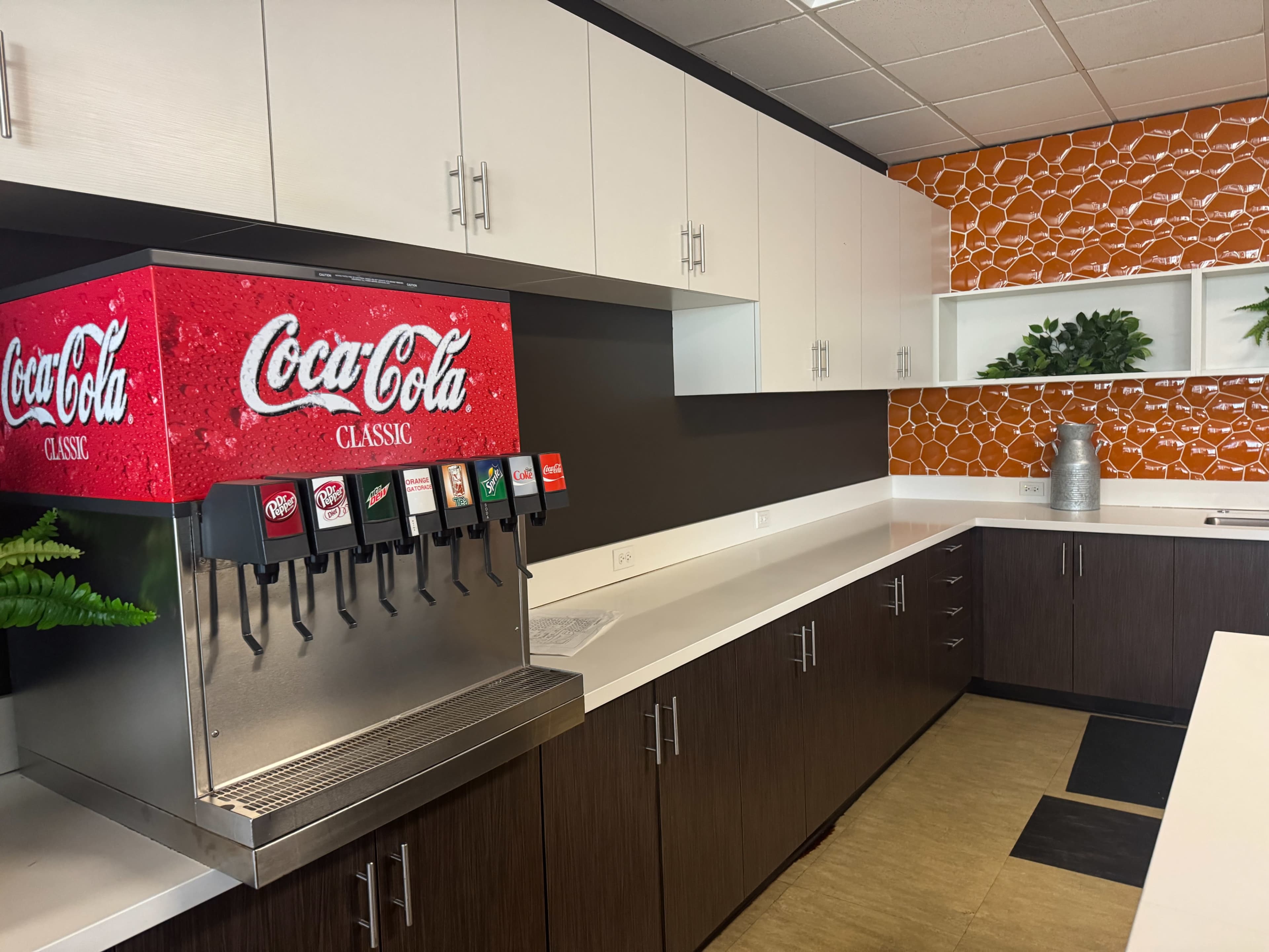 The image shows a drink station with a Coca-Cola soda dispenser, situated against a backdrop of white cabinetry and an orange tiled wall in a modern kitchen area.