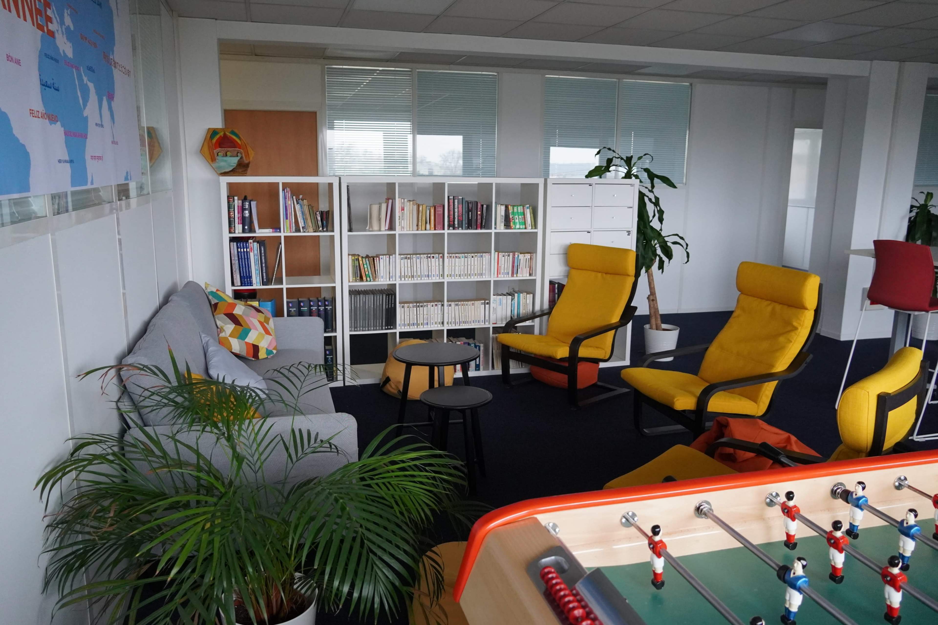 The image shows a modern office lounge area with a gray sofa, two yellow armchairs, a coffee table, and a foosball table, surrounded by shelves of books and plants.