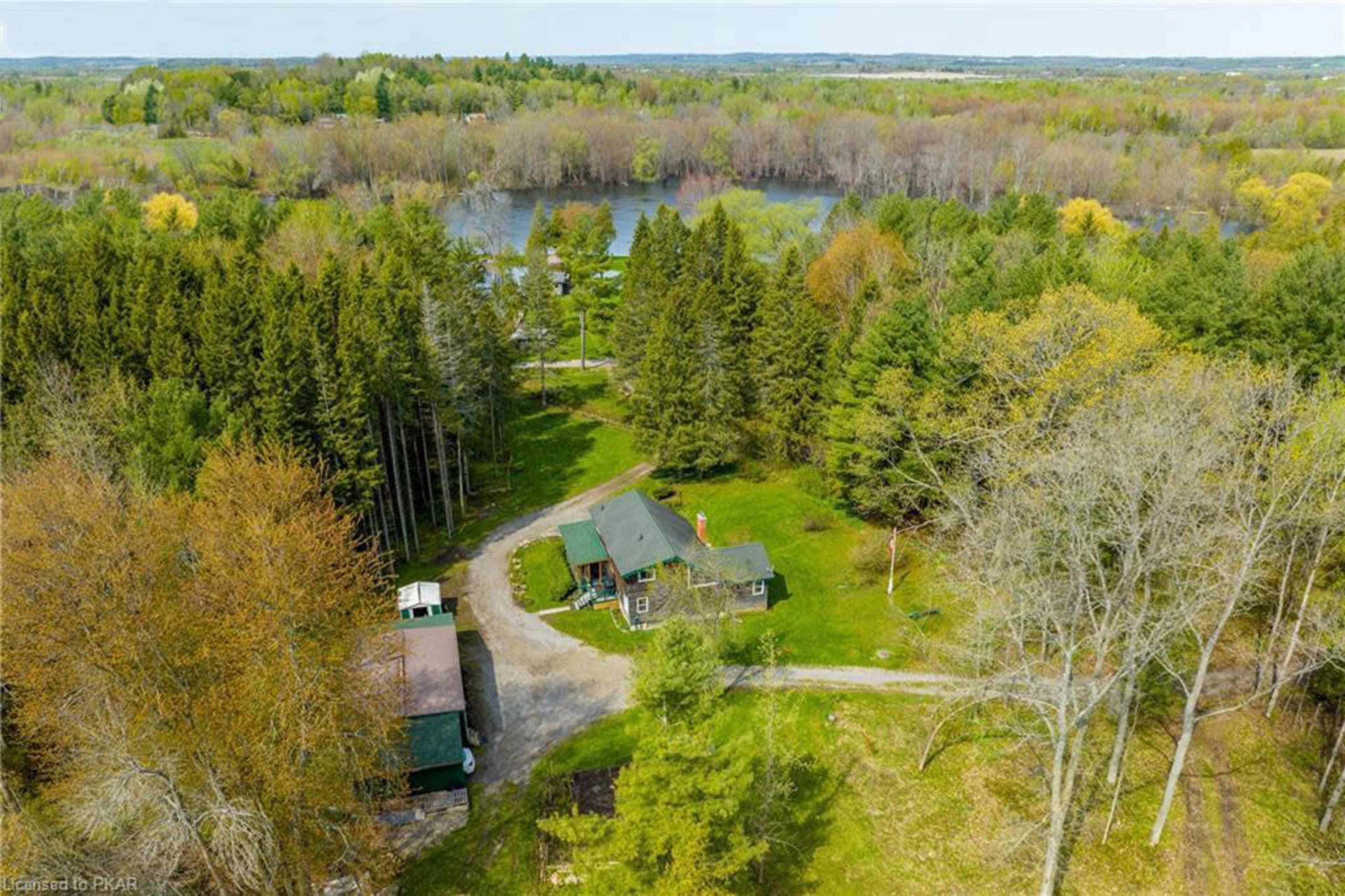 An aerial view shows a green-roofed house surrounded by trees and a winding driveway, near a body of water.