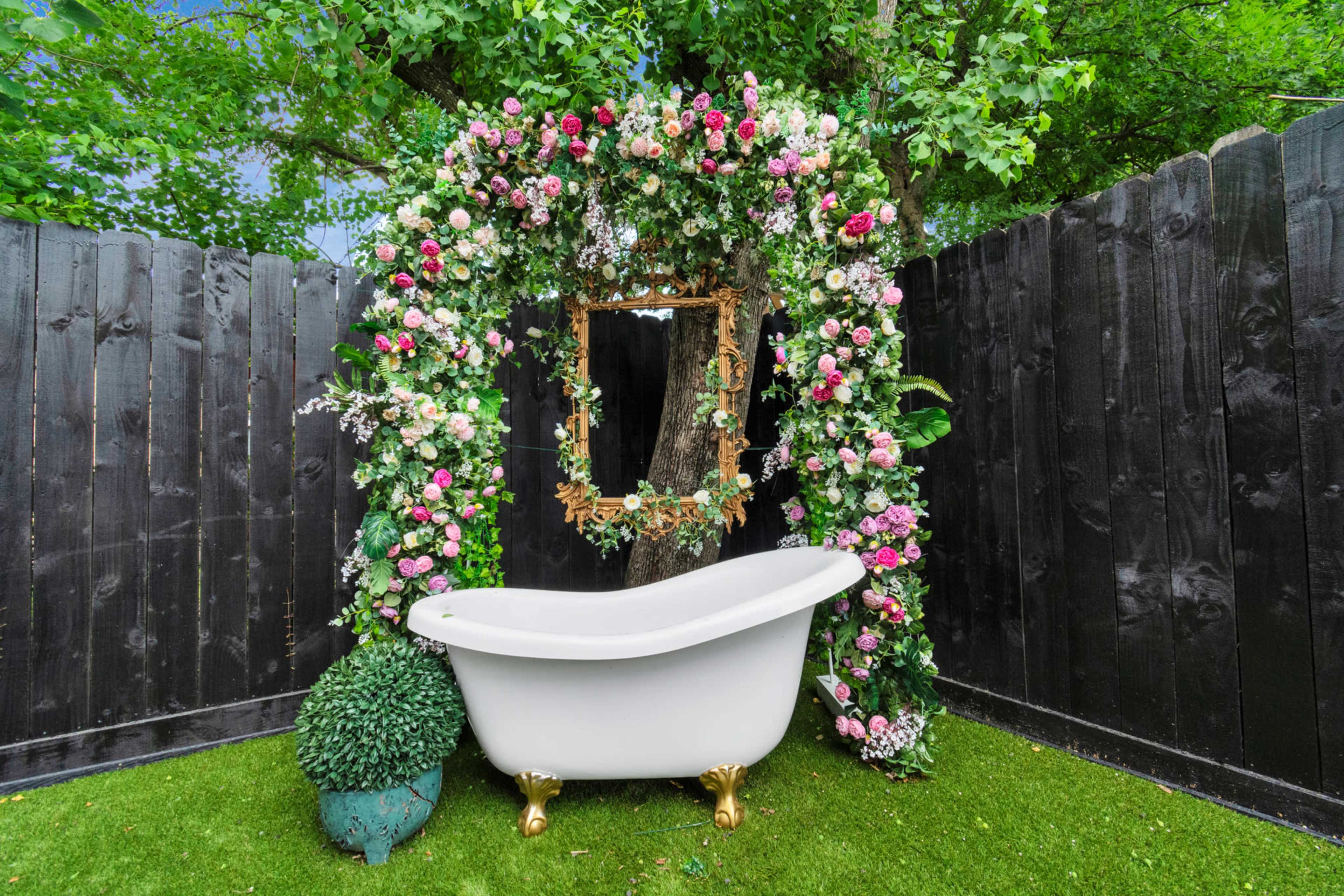 A white clawfoot bathtub is placed under a floral arch set against a backdrop of black wooden fencing and green foliage.