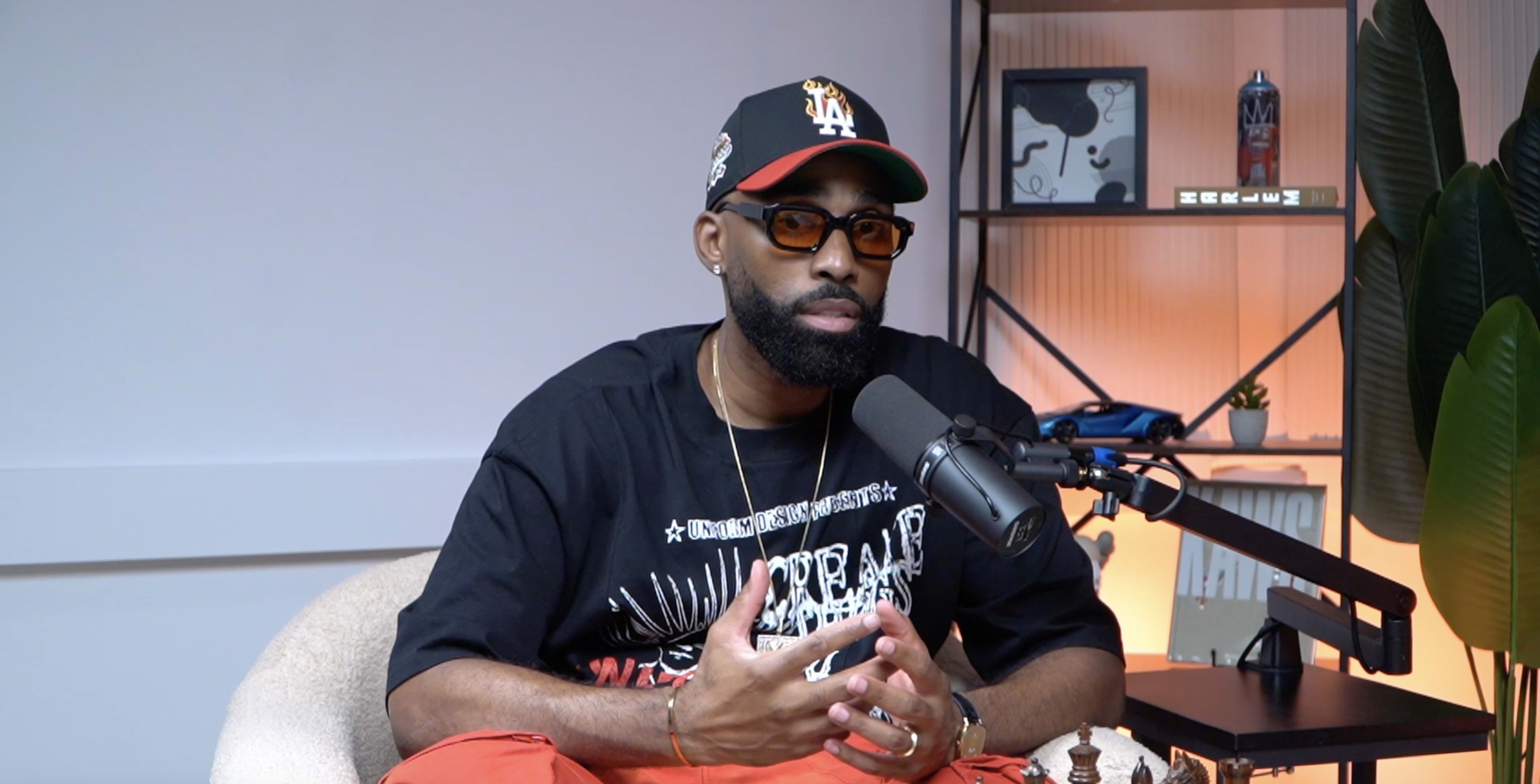 A man with a beard and sunglasses sits in a studio, gesturing while speaking into a microphone, with a chessboard and decorative items in the background.