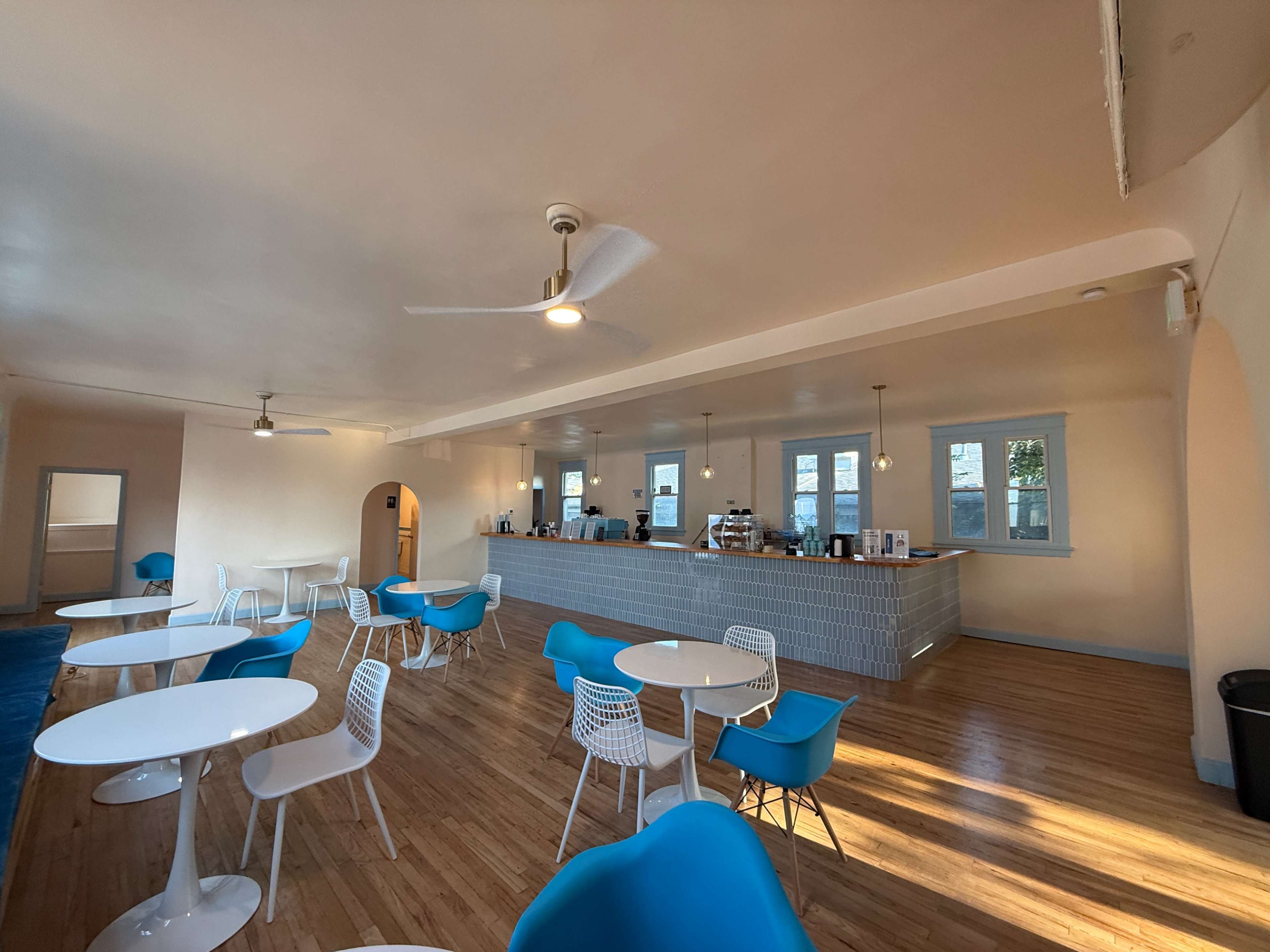 The image shows a spacious café with blue chairs and white tables, featuring a long counter against the wall where various coffee and food items are likely served.