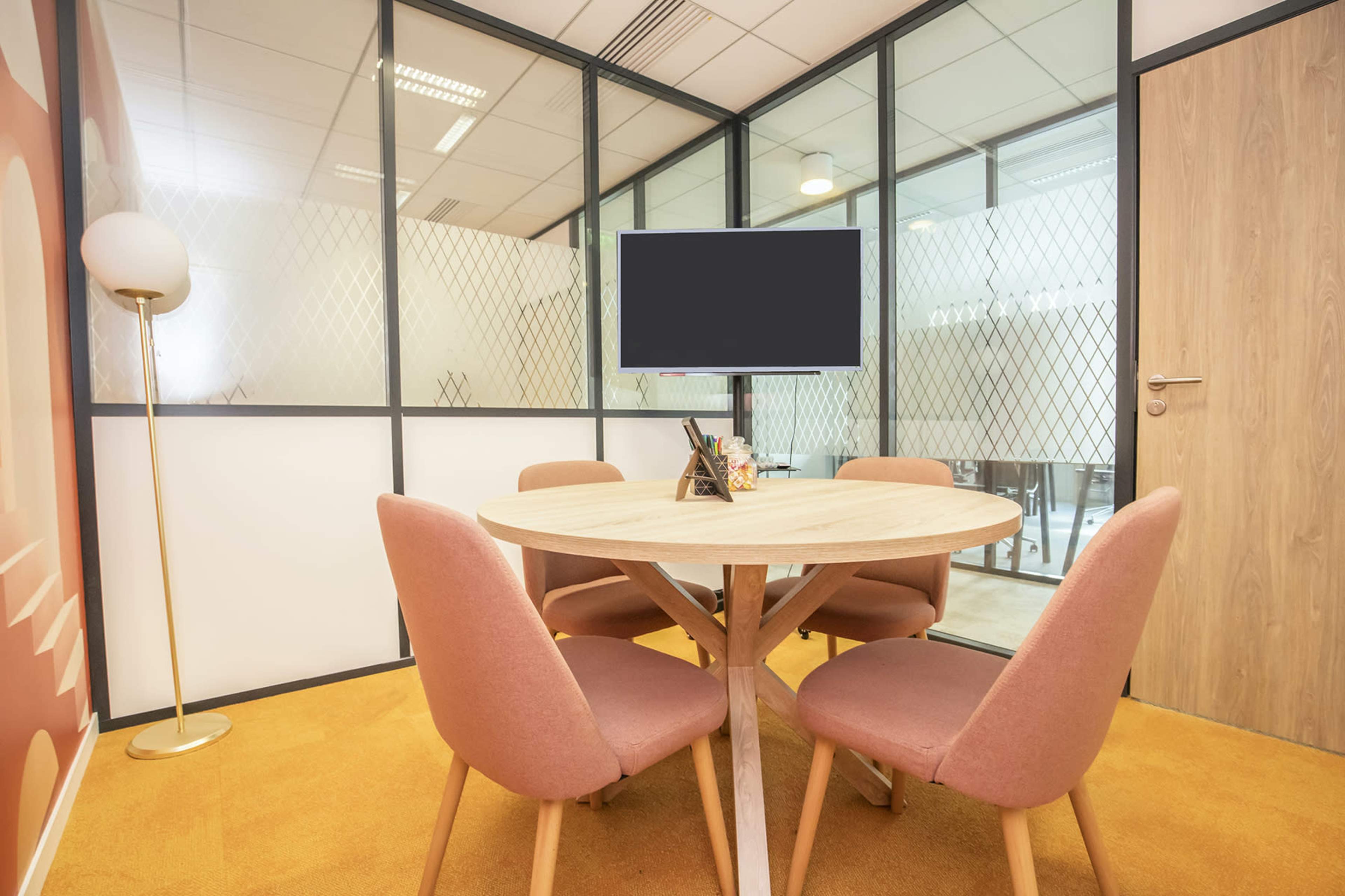 A round wooden table surrounded by four pink chairs is situated in a glass-enclosed meeting room.