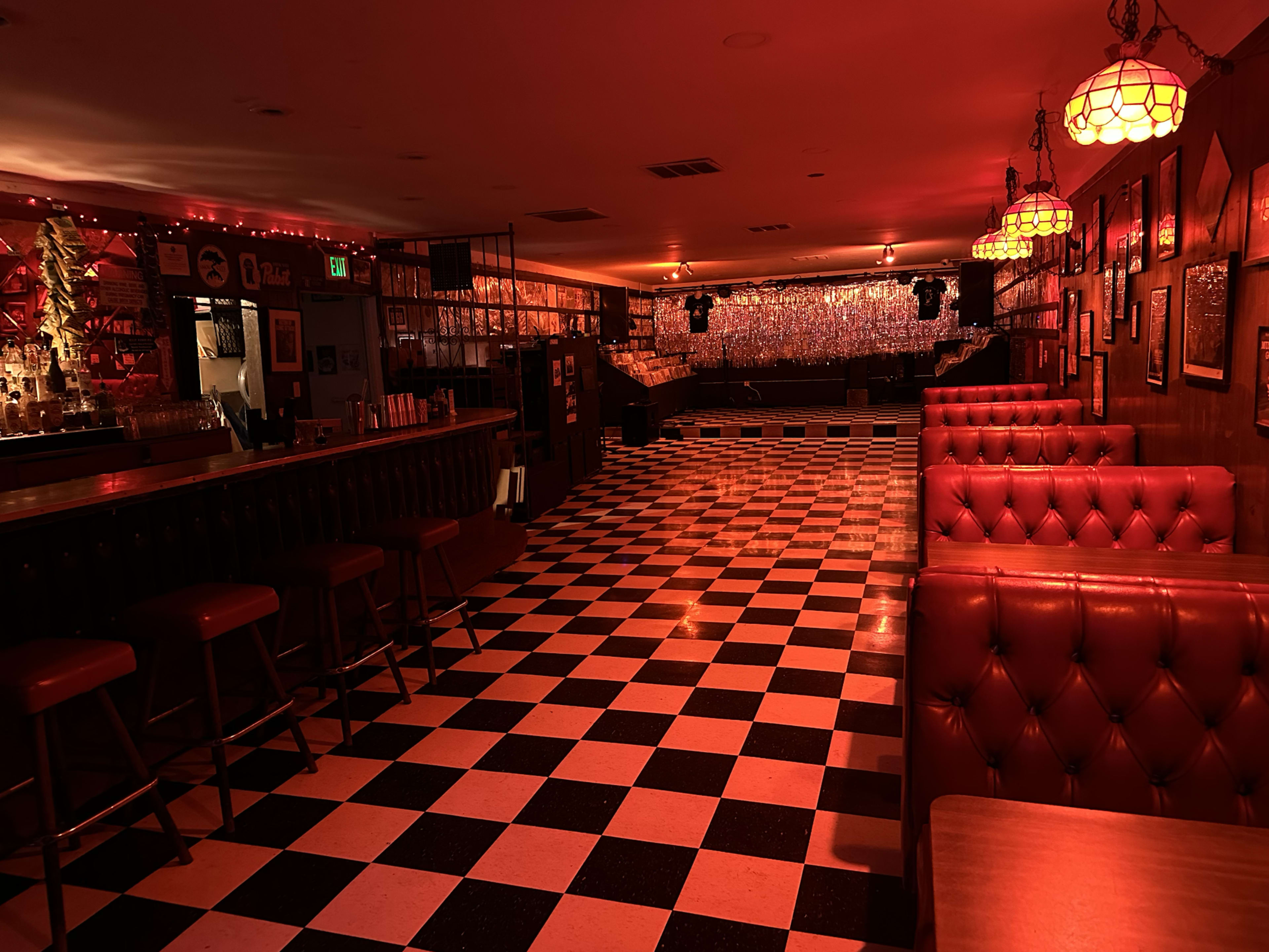 The image shows an empty bar with a checkerboard floor, red lighting, and tufted leather seating, leading to a stage area in the background.
