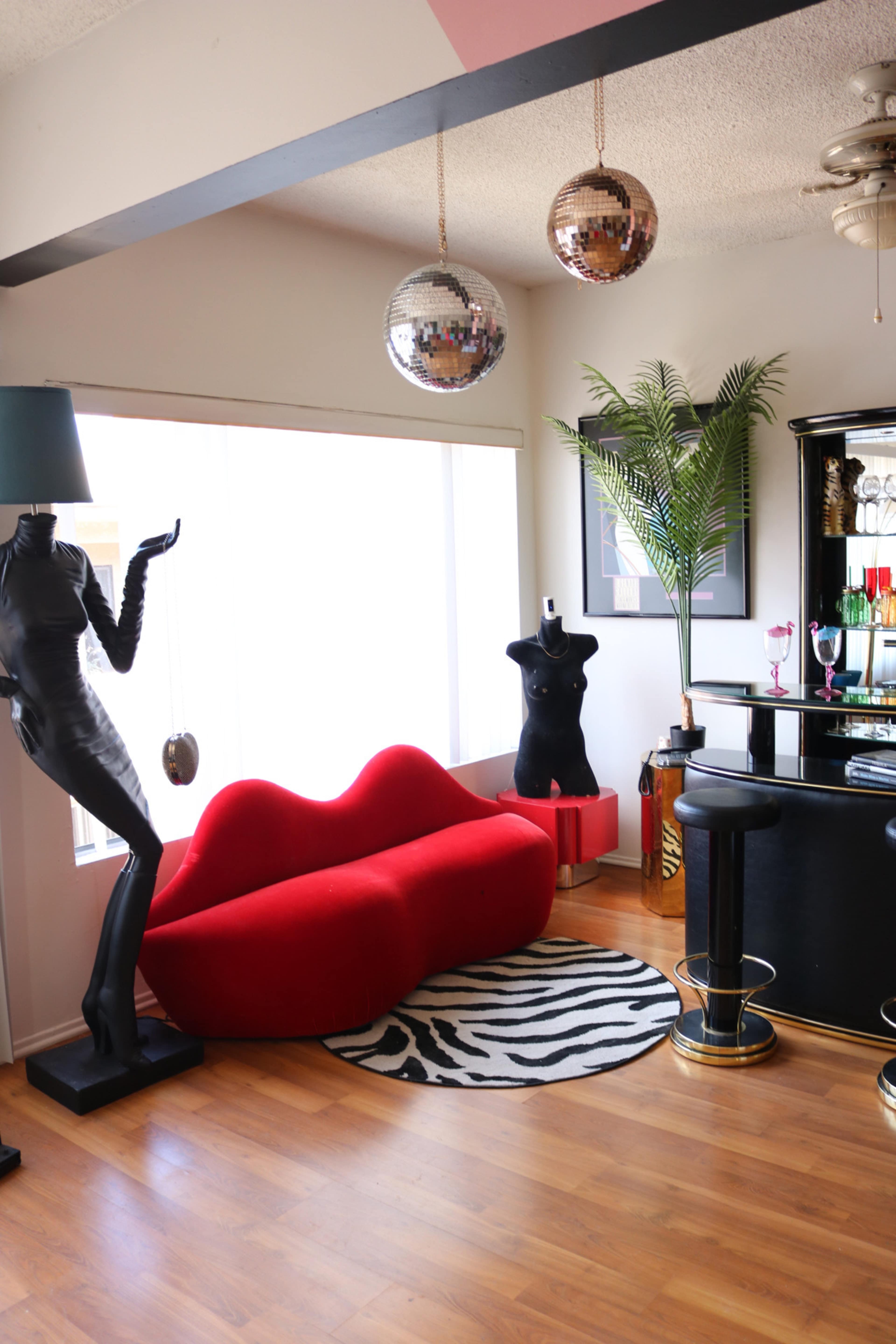 A stylish living room with a red curved sofa, a zebra-patterned rug, and a bar area featuring a black counter, decorative plants, and disco balls hanging from the ceiling.