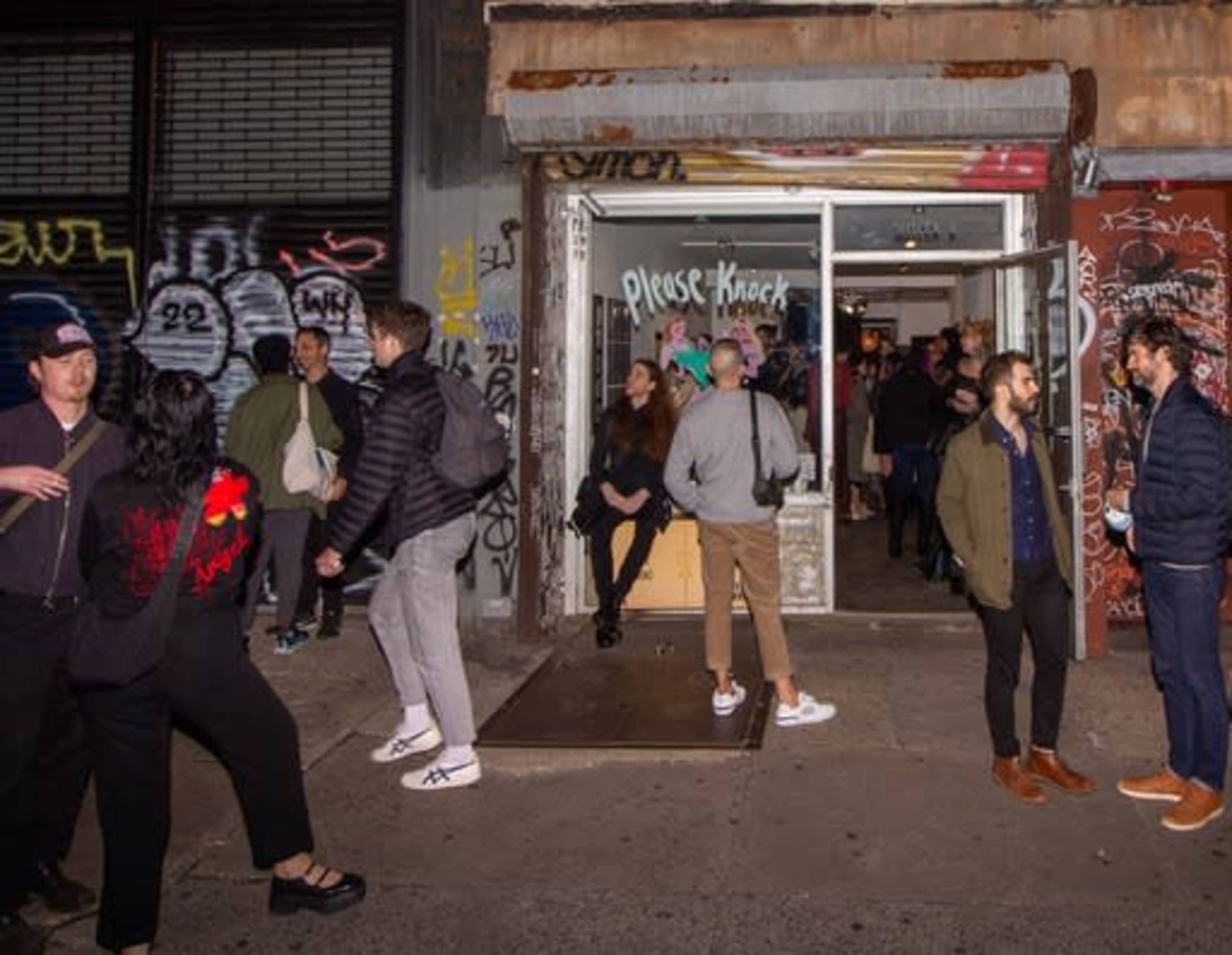 A crowd of people gathers outside a graffiti-covered storefront, some entering while others chat near the entrance.