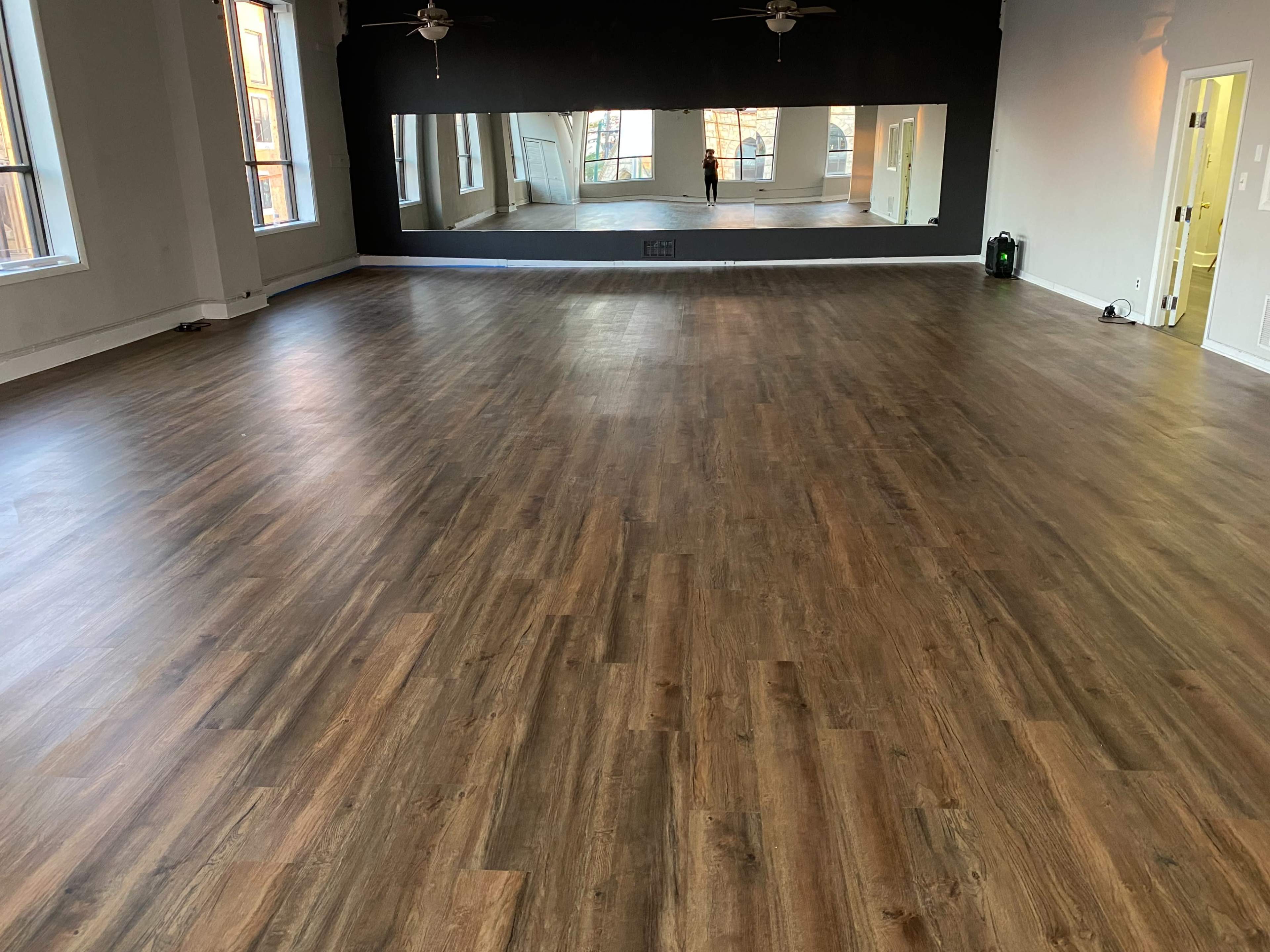 An empty room with wooden flooring, large windows, and a mirror on one wall.