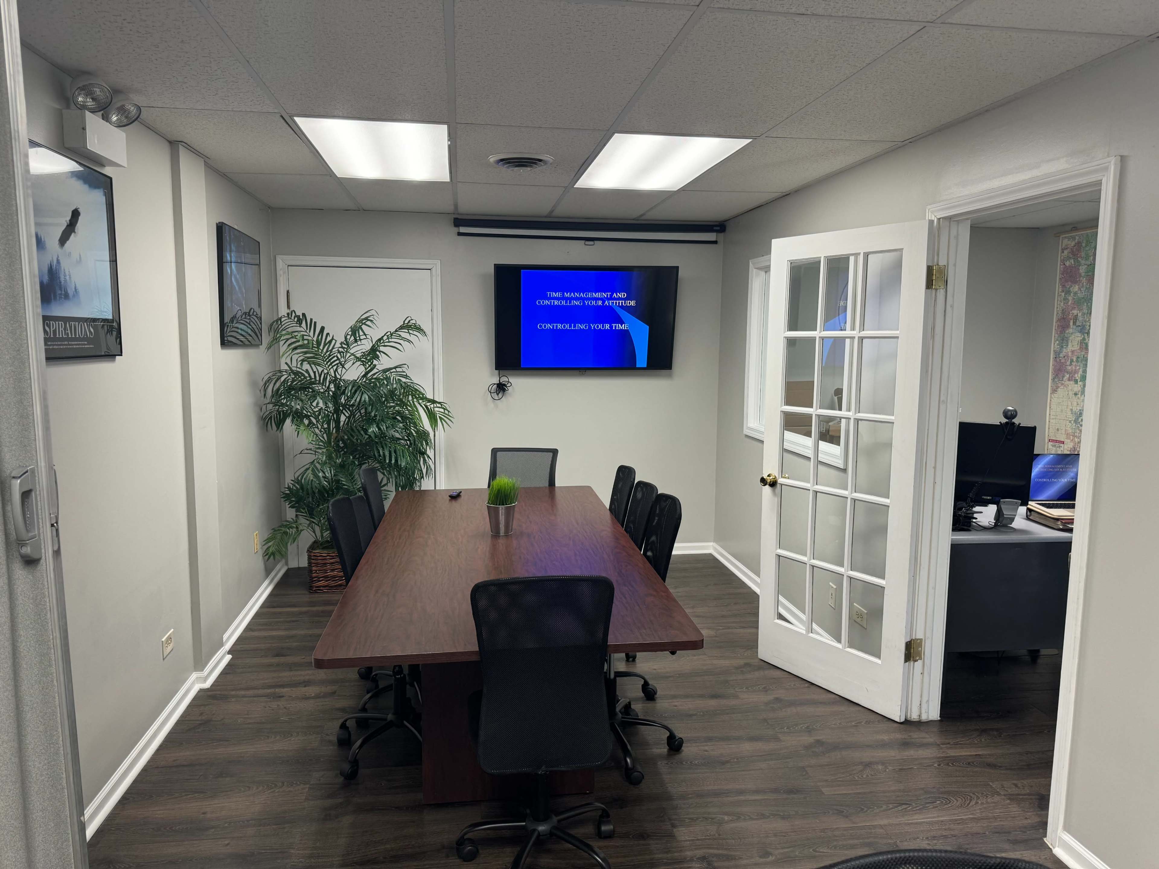 A conference room features a long wooden table surrounded by black chairs, with a television screen displaying text on one wall and an open door leading to another office.