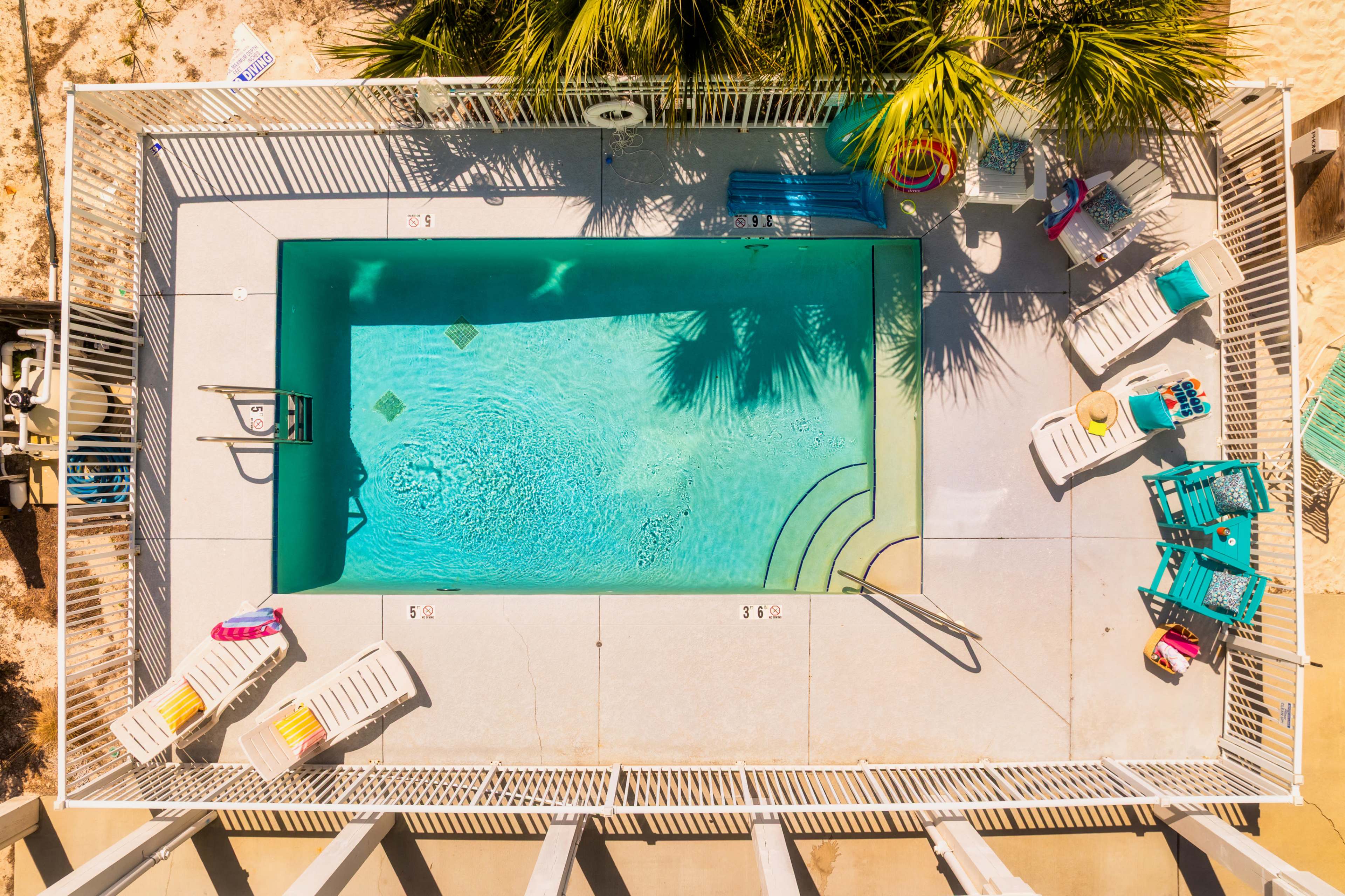 The image shows an aerial view of a rectangular swimming pool surrounded by lounge chairs and tropical plants.