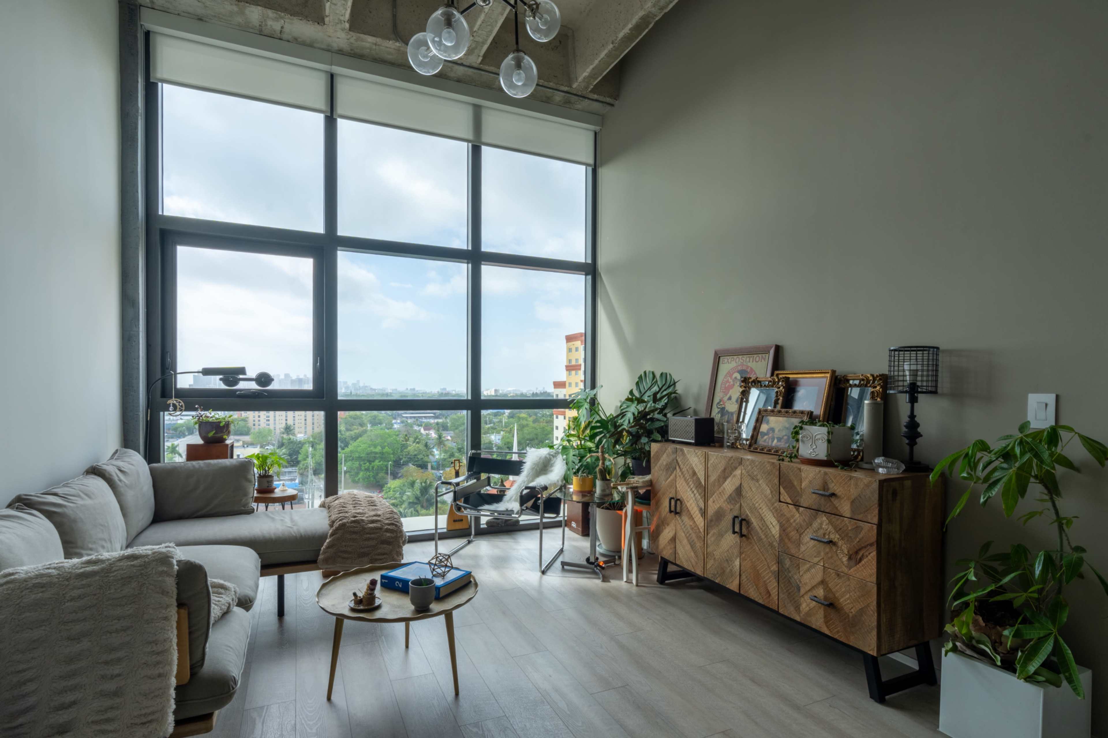 The image shows a modern living room with large windows, a light-colored sofa, a wooden sideboard, and various plants.