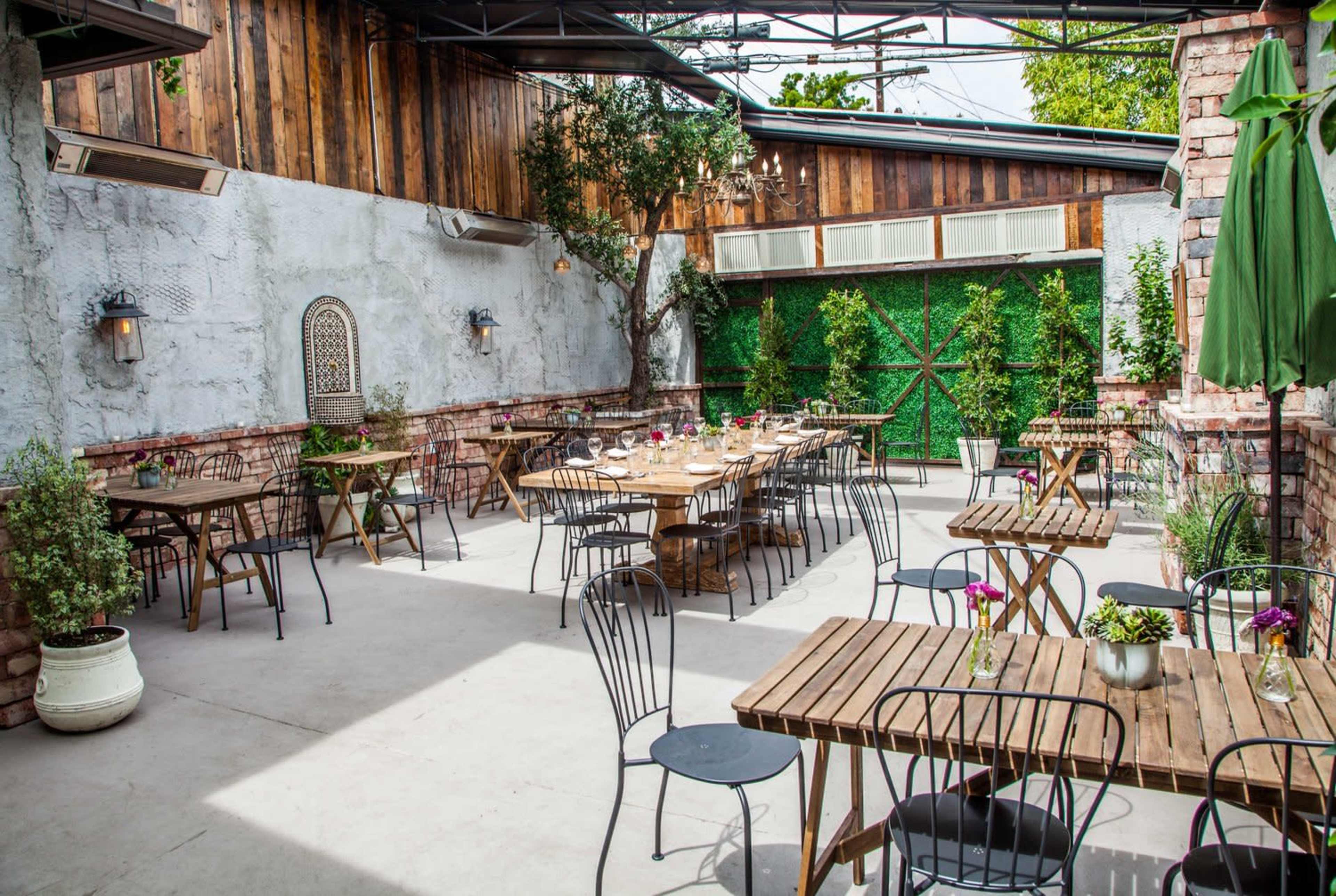 An outdoor restaurant patio with wooden tables and chairs arranged neatly, surrounded by greenery and rustic brick walls.