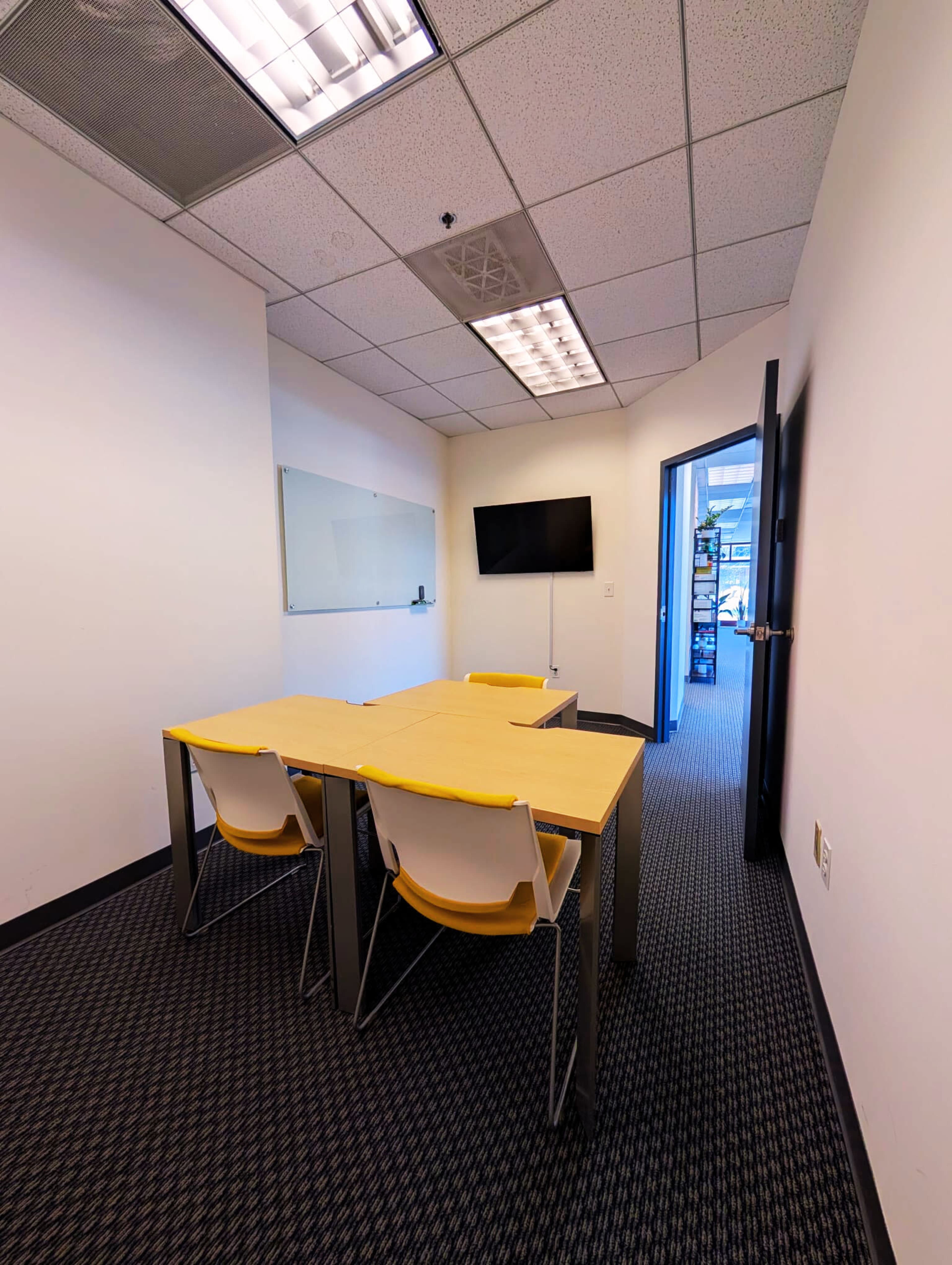 The image shows a small conference room with a rectangular table, four chairs, a wall-mounted TV, and a whiteboard.
