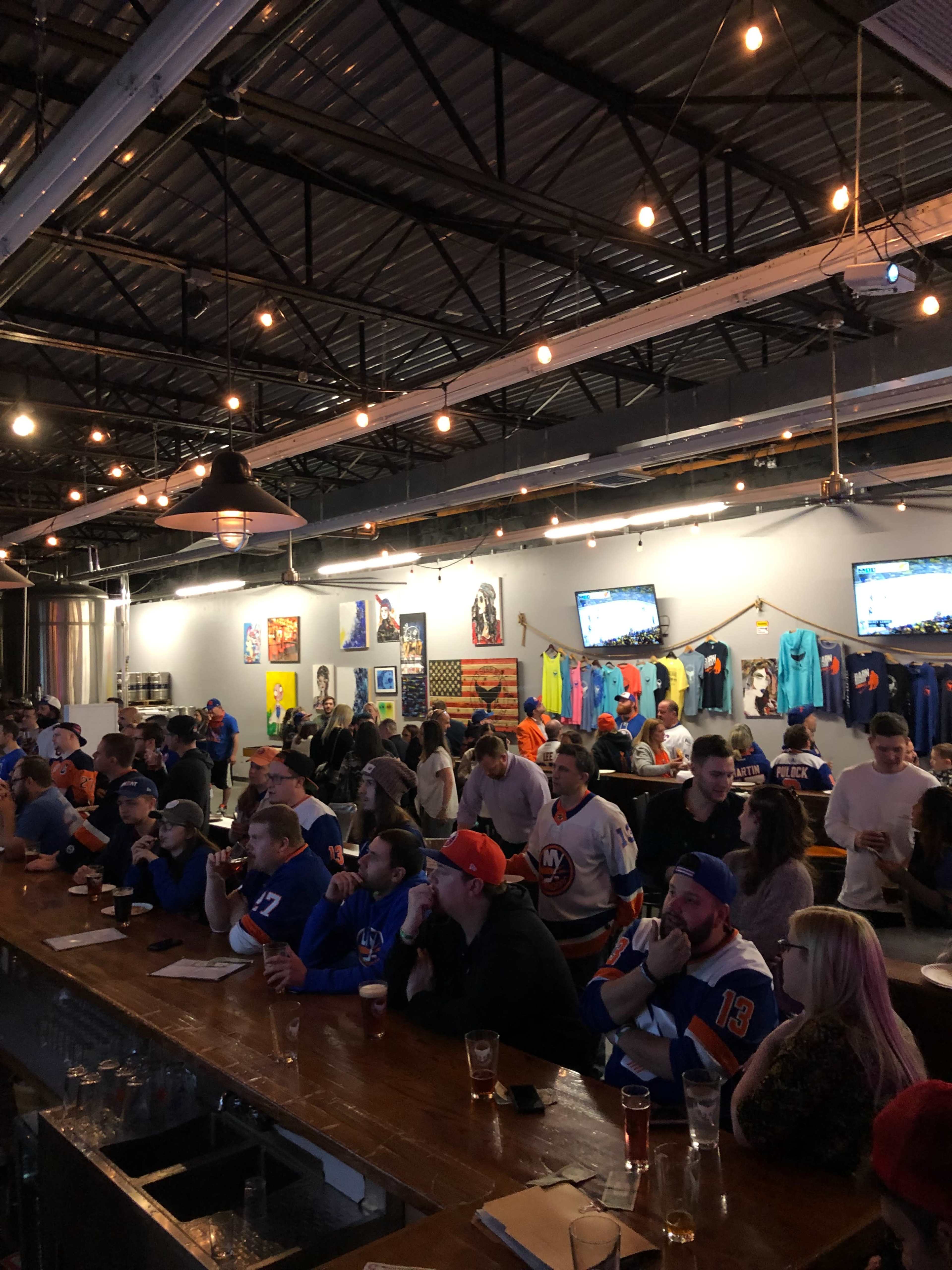 A crowd of people wearing hockey jerseys gathers around a long bar in a lively sports bar, with multiple TVs showing games in the background.