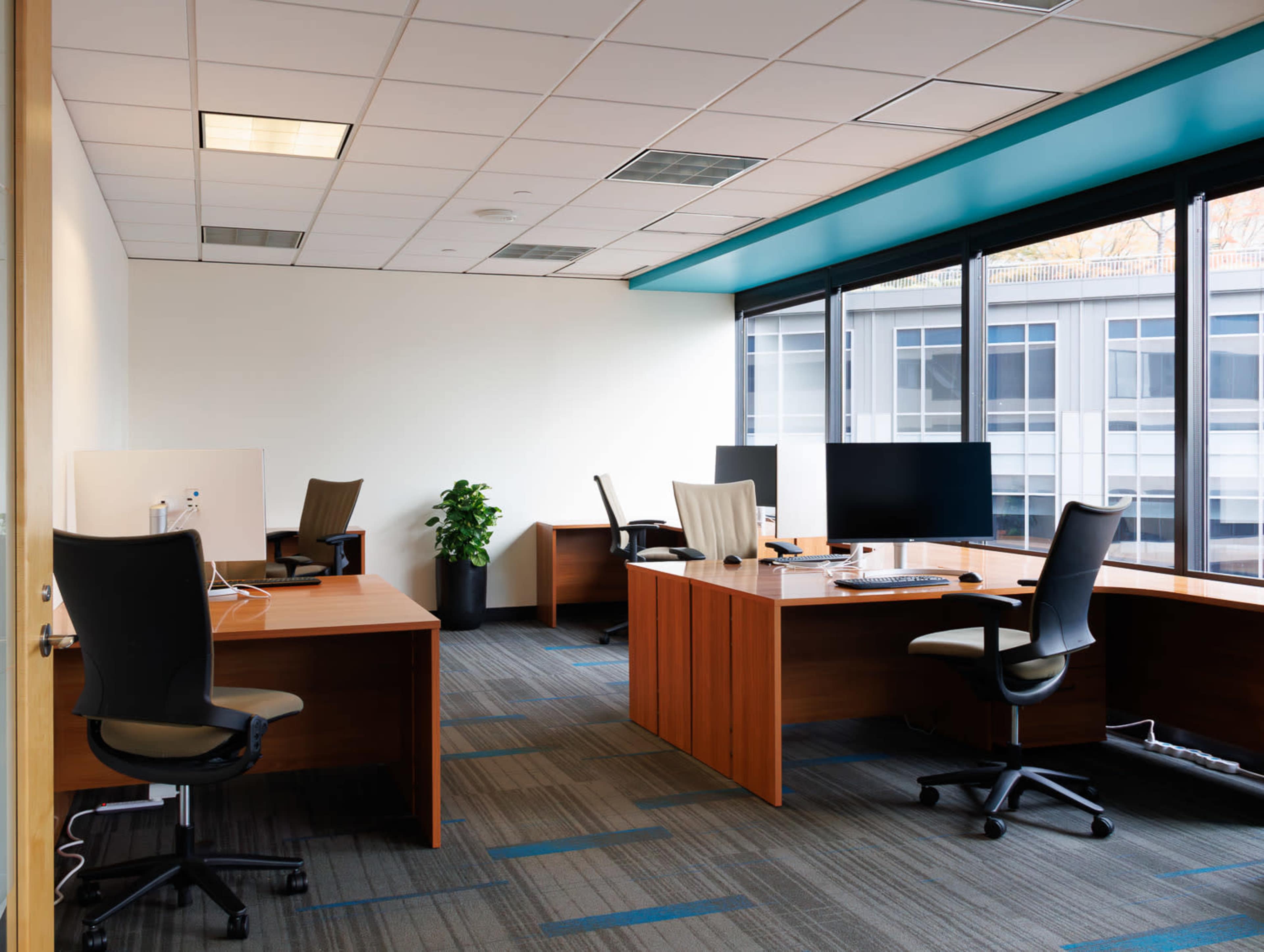 The image shows a modern office space with four wooden desks, each equipped with office chairs and computer monitors, illuminated by overhead lighting and natural light from large windows.