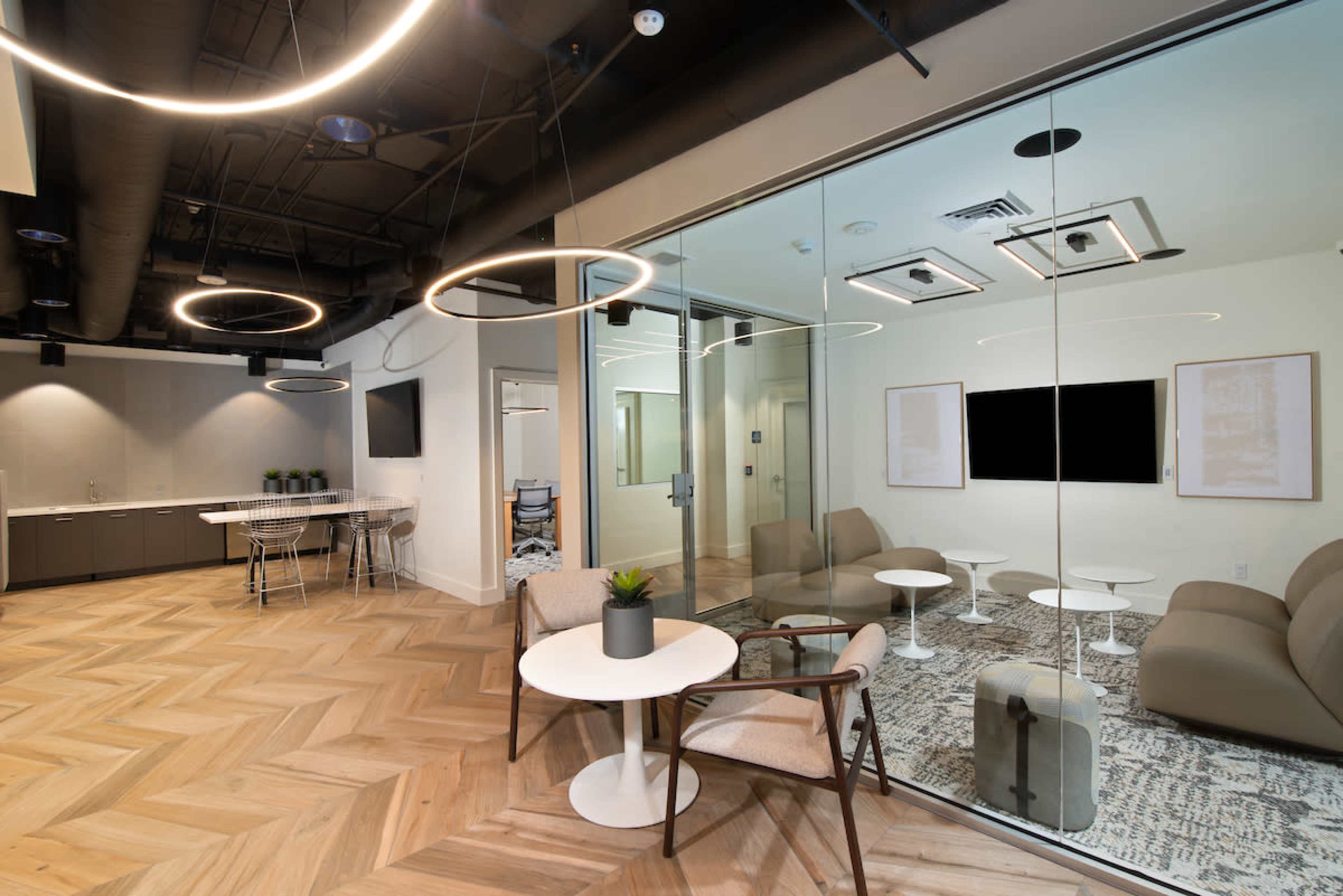 The image shows a modern office meeting space with a glass wall, featuring a round table and chairs, soft seating areas, and a kitchenette in the background.