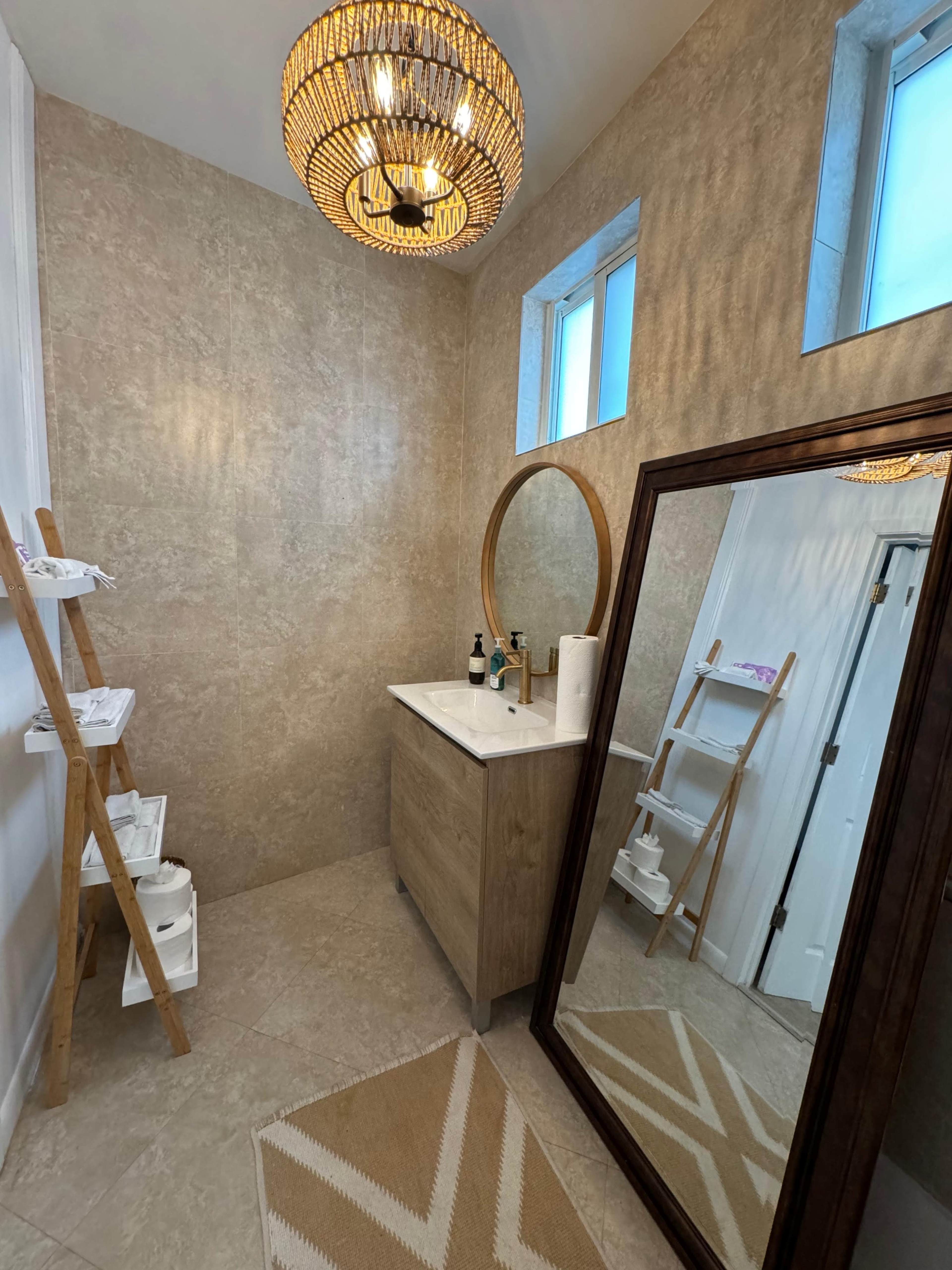 A modern bathroom features a wooden vanity with a round mirror, a large floor mirror, a decorative light fixture, and a ladder shelf against textured walls.
