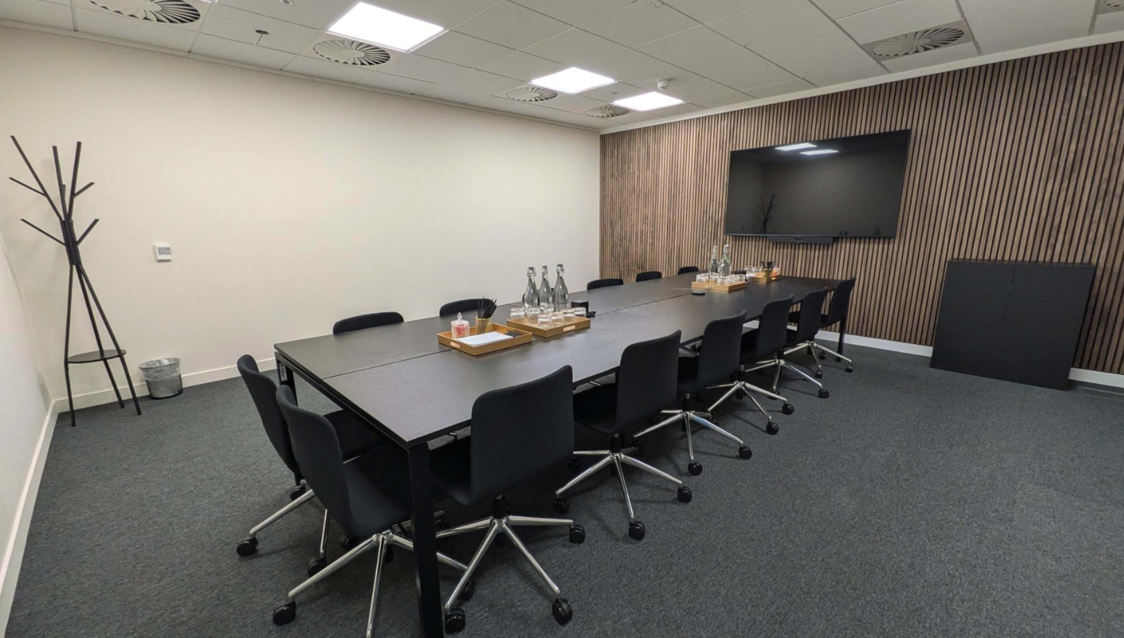 The image shows a modern conference room featuring a long rectangular table surrounded by black rolling chairs, with a wooden accent wall and a large wall-mounted screen.