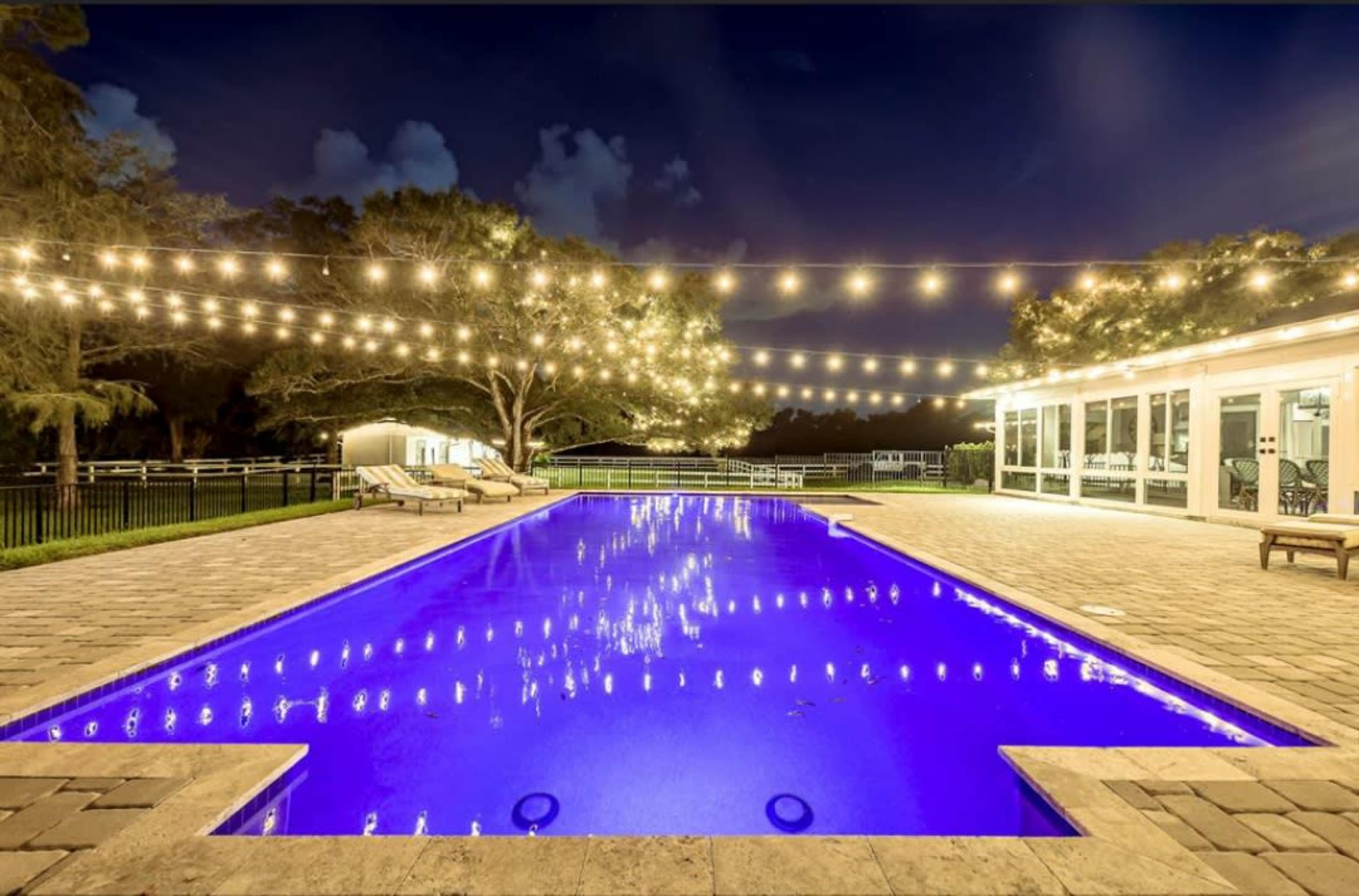 A well-lit rectangular pool surrounded by lounge chairs, with string lights overhead and a house visible in the background at night.