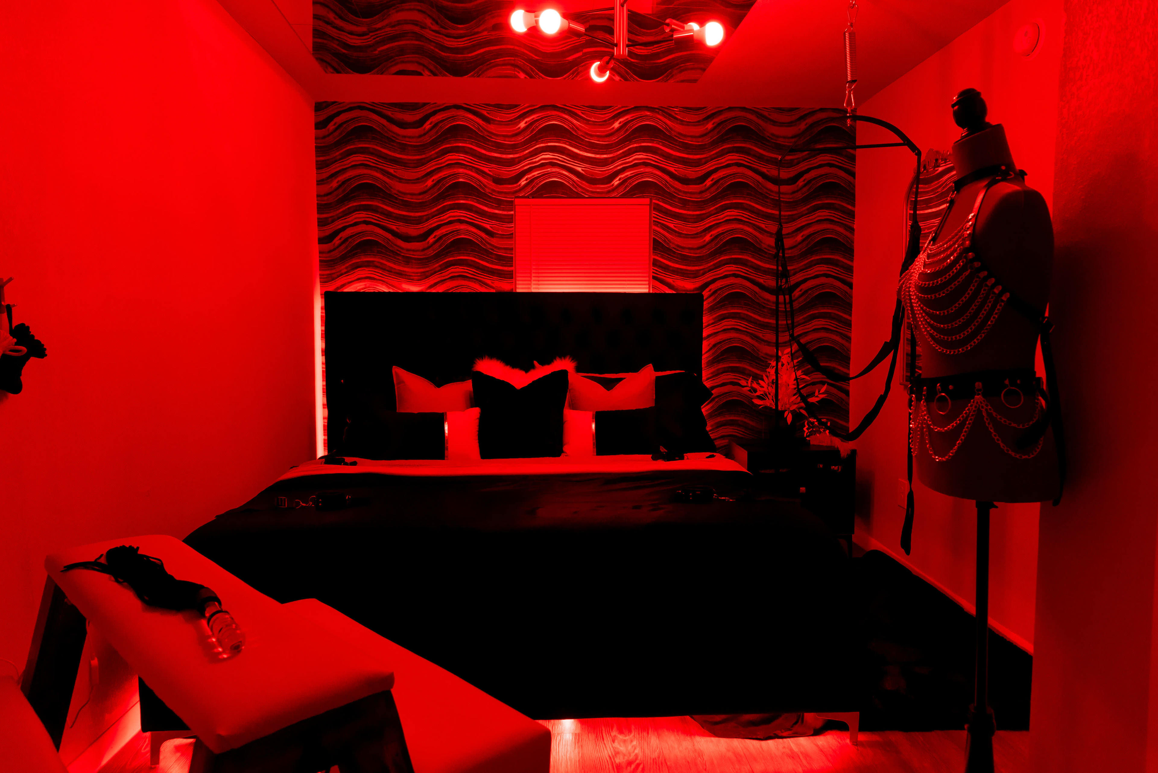 A dimly lit bedroom featuring a large bed with dark bedding, decorative pillows, and a mannequin wearing chains, against a red-toned wall.