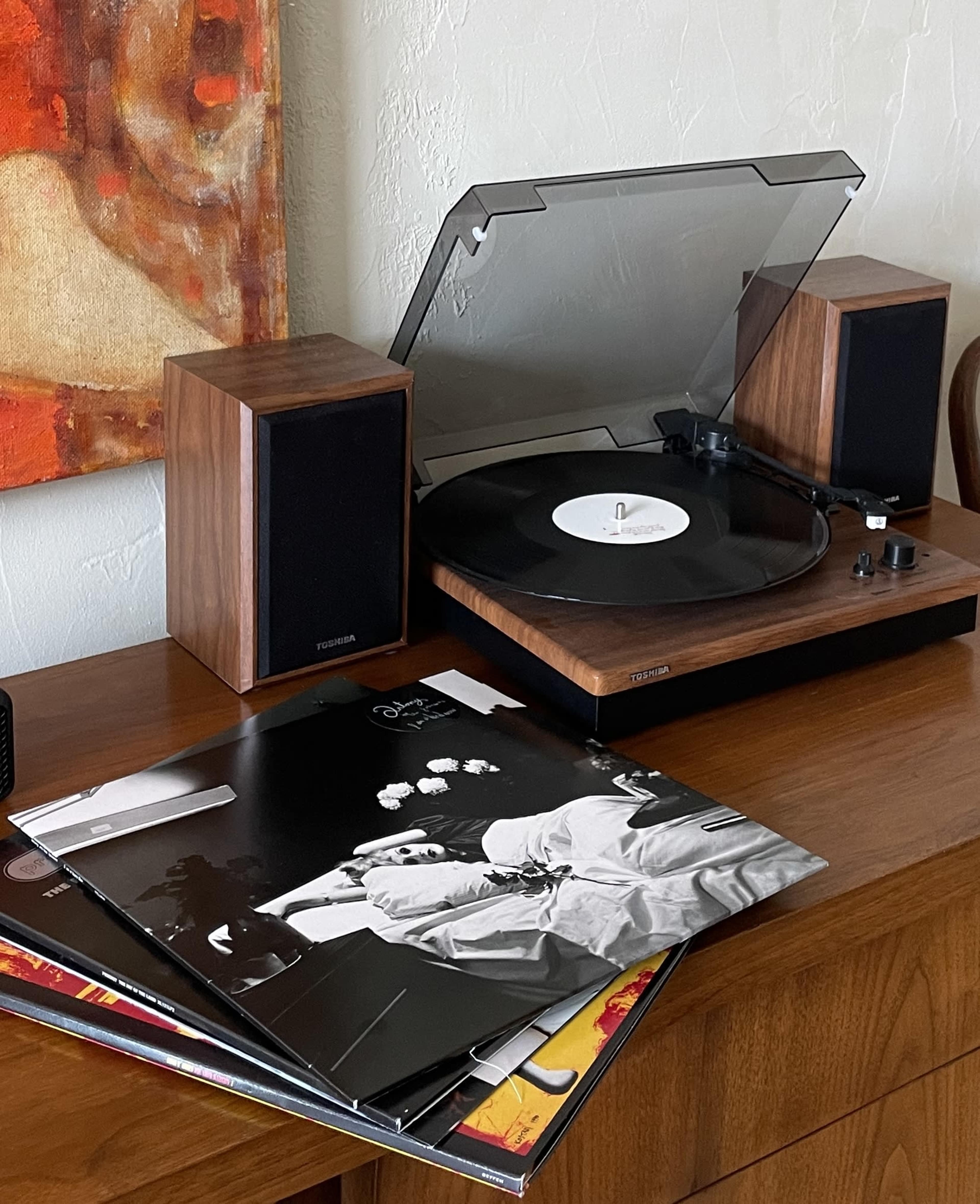 A turntable with speakers and several vinyl records is placed on a wooden surface next to a colorful painting.