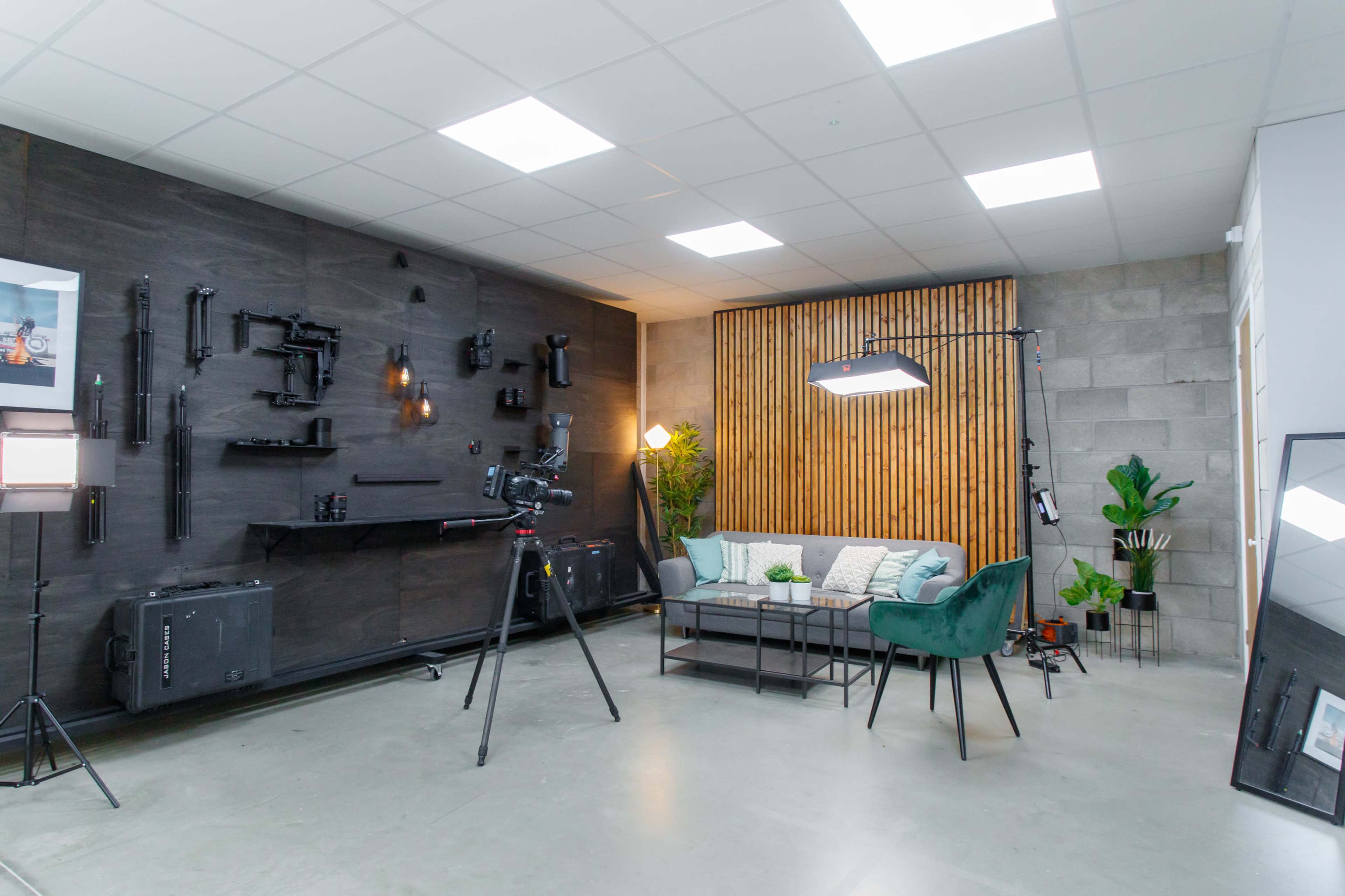 A modern studio setup with a couch, decorative plants, and a camera on a tripod against a backdrop of wooden slats and various equipment mounted on the wall.
