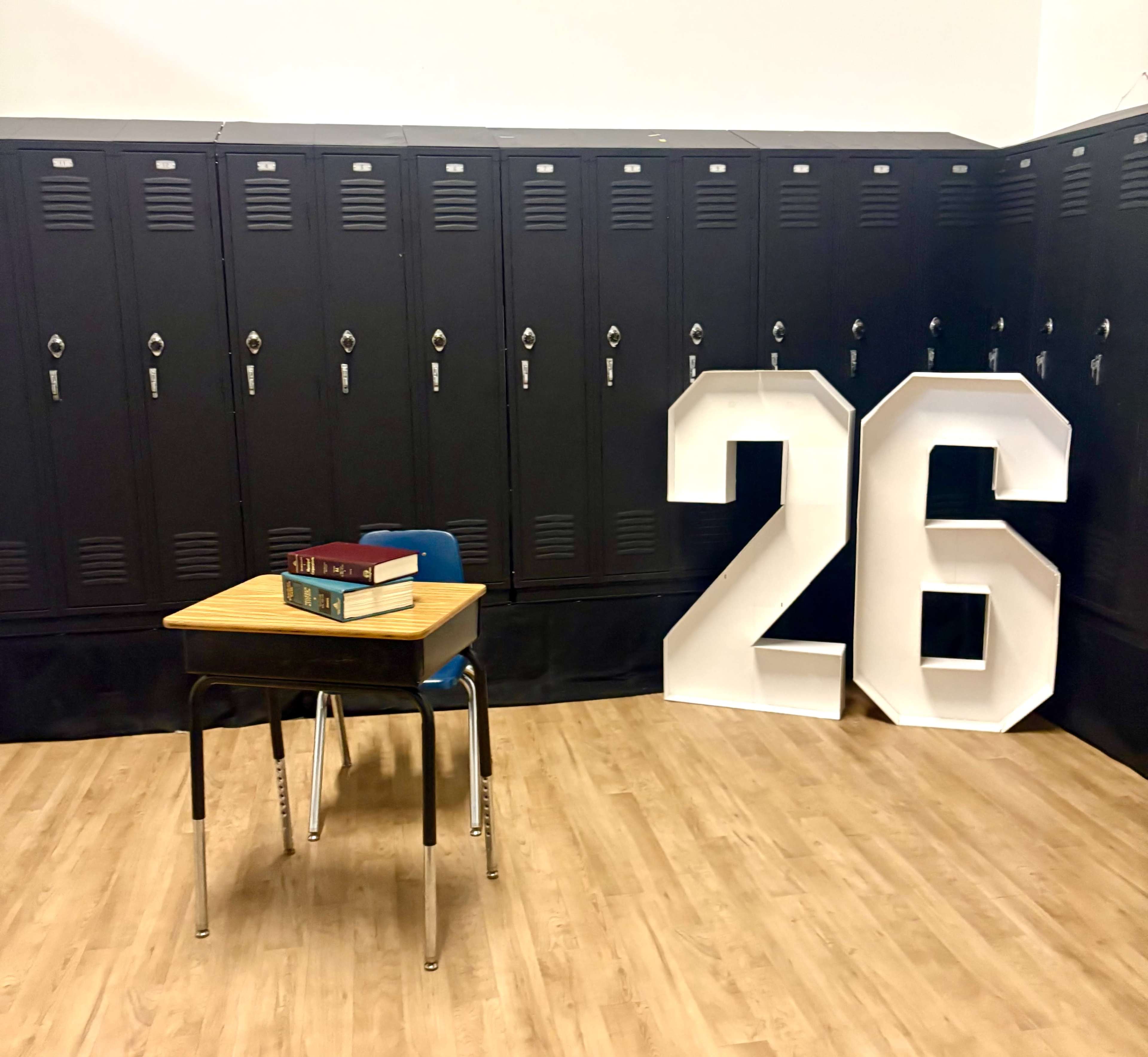 The image shows a classroom setting with a small desk and chair in front of large white block numbers "26," alongside a row of black lockers in the background.