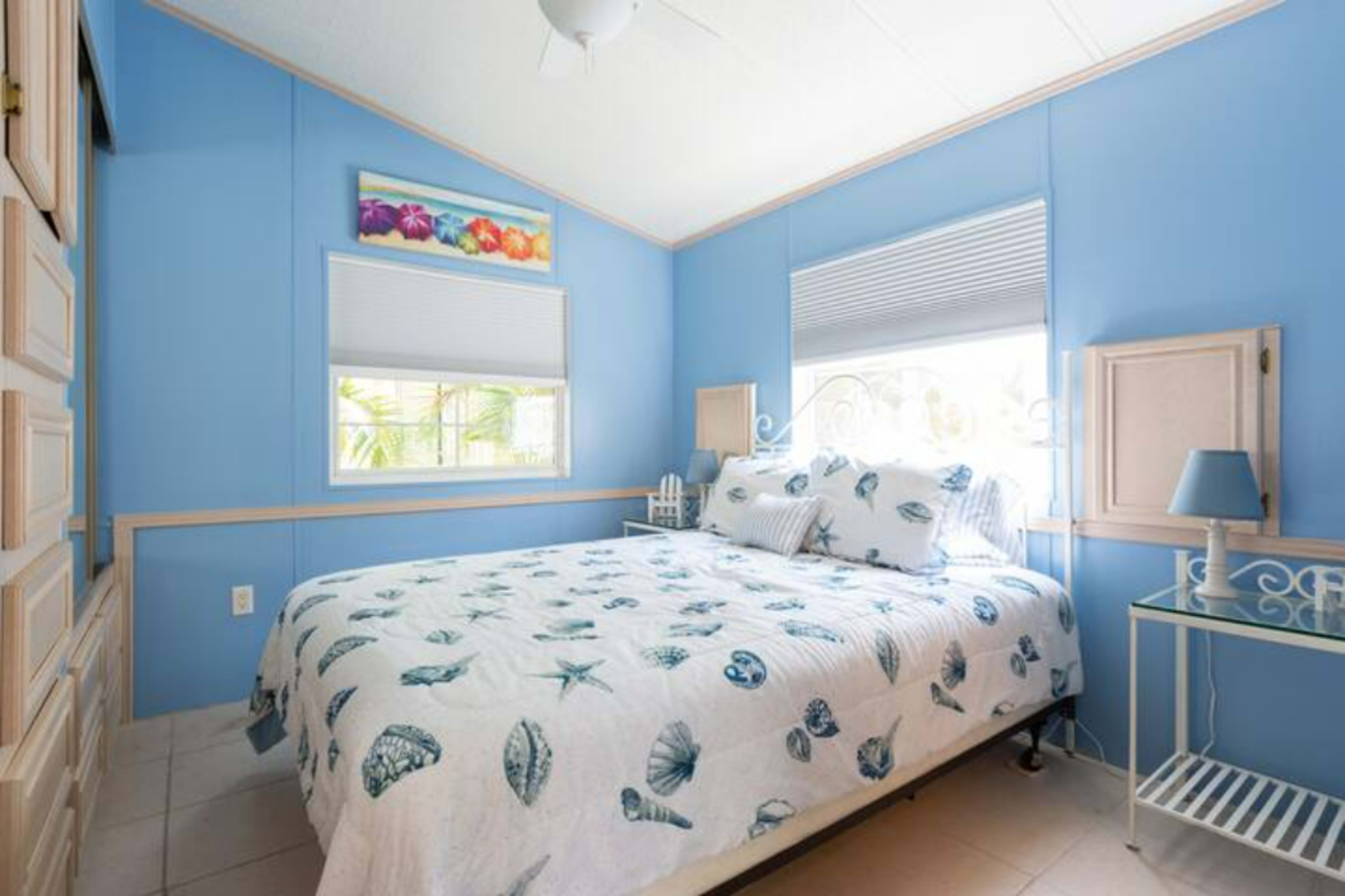 A blue-walled bedroom with a bed adorned with a seashell-patterned quilt, a nightstand with a lamp, and a window allowing natural light.