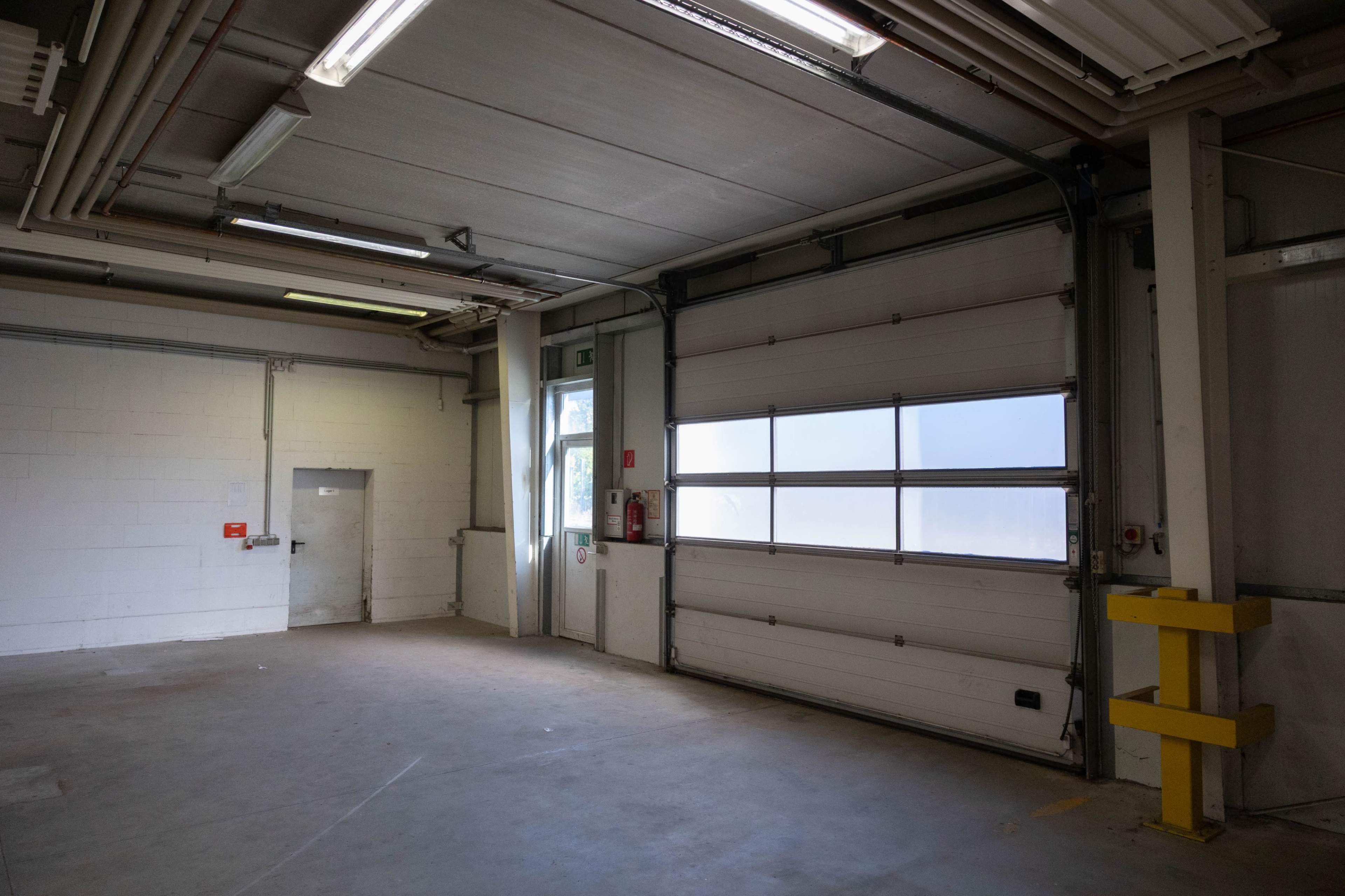 The image shows an empty industrial space with a large closed garage door, a side door, and exposed pipes on the ceiling.