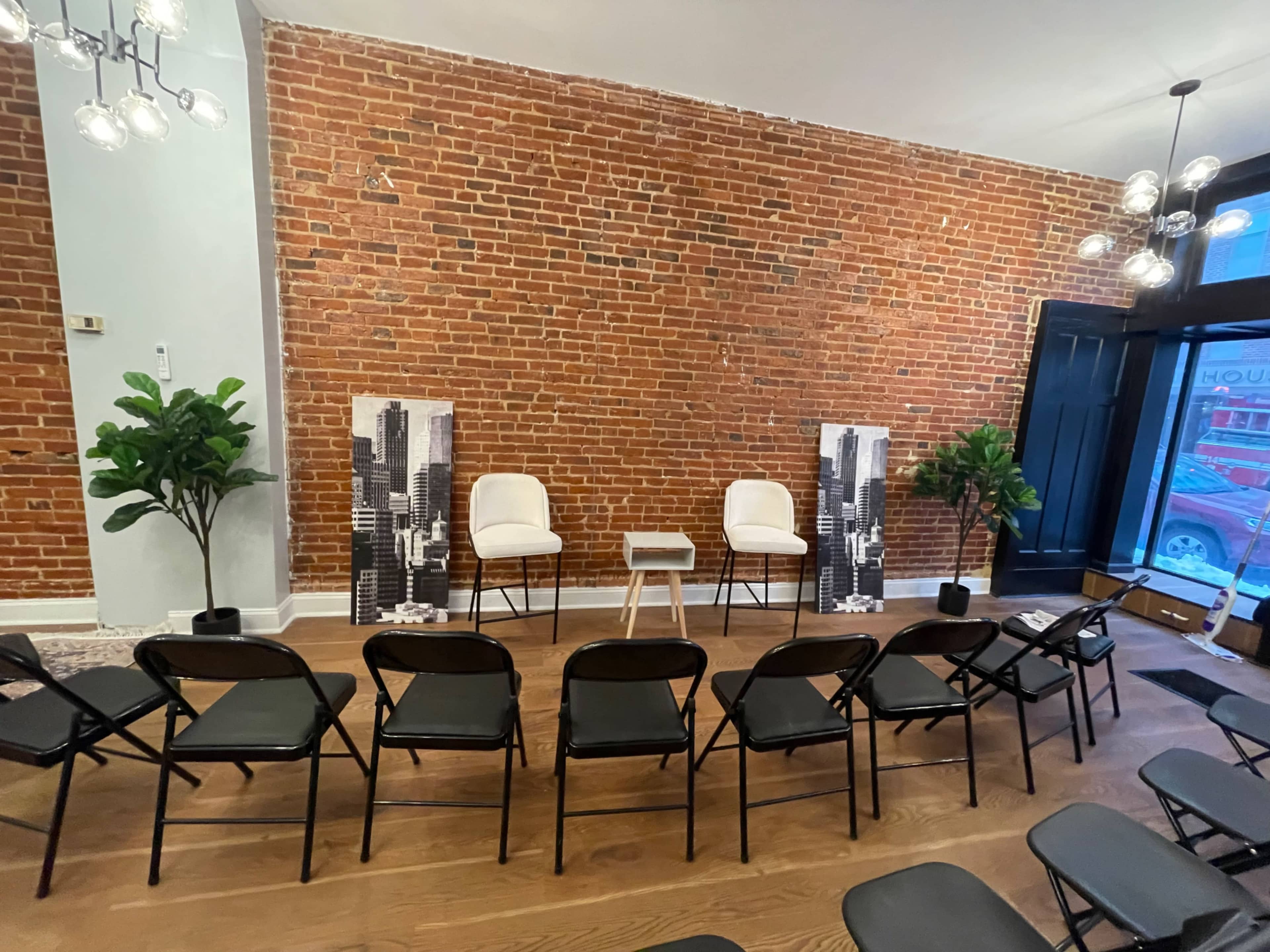 A set of empty black folding chairs faces a small table and two white chairs in front of a brick wall, flanked by decorative plants.