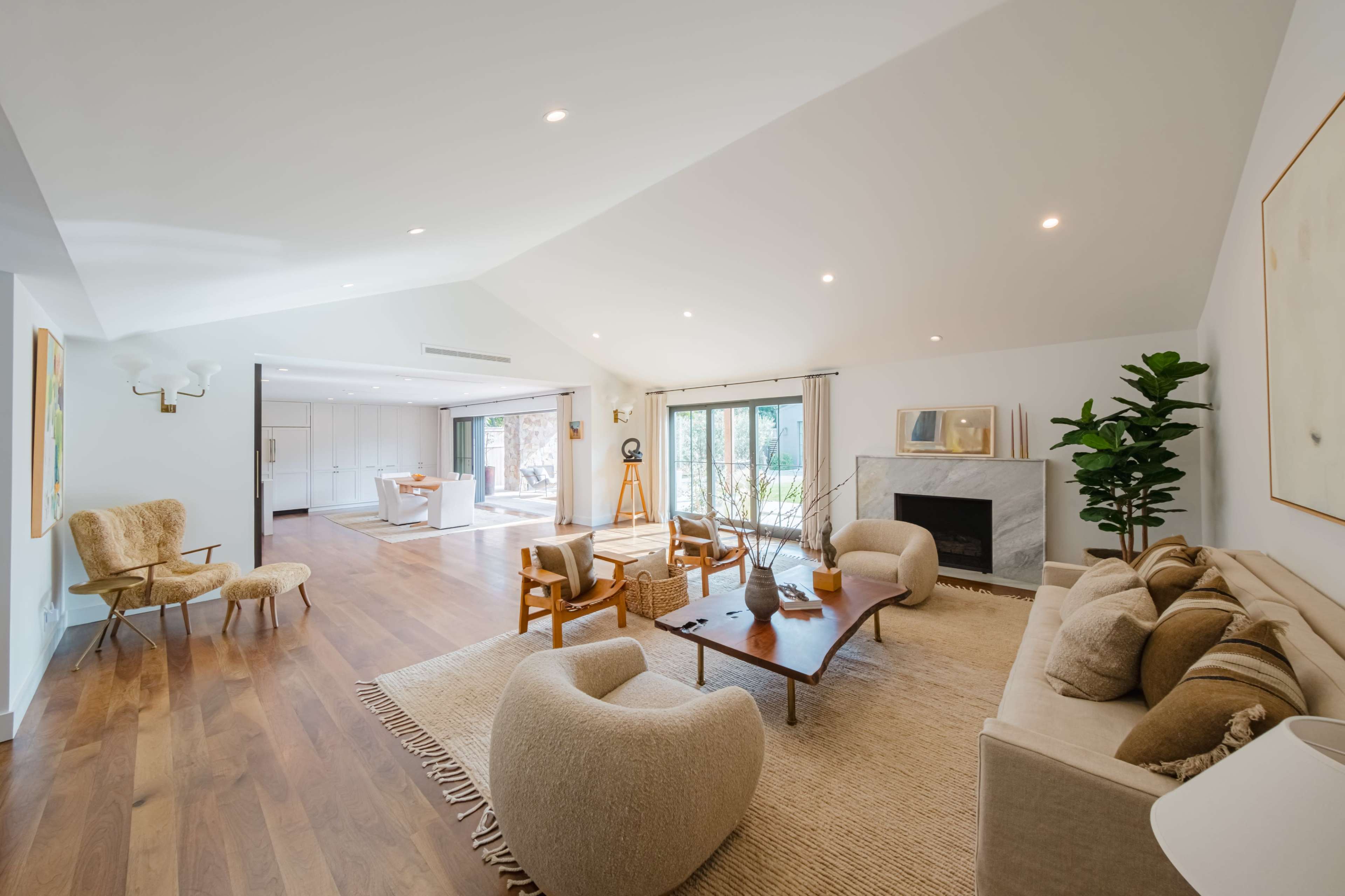 The living room features a sofa and armchairs arranged around a wooden coffee table, with large windows providing views of an adjoining kitchen area.