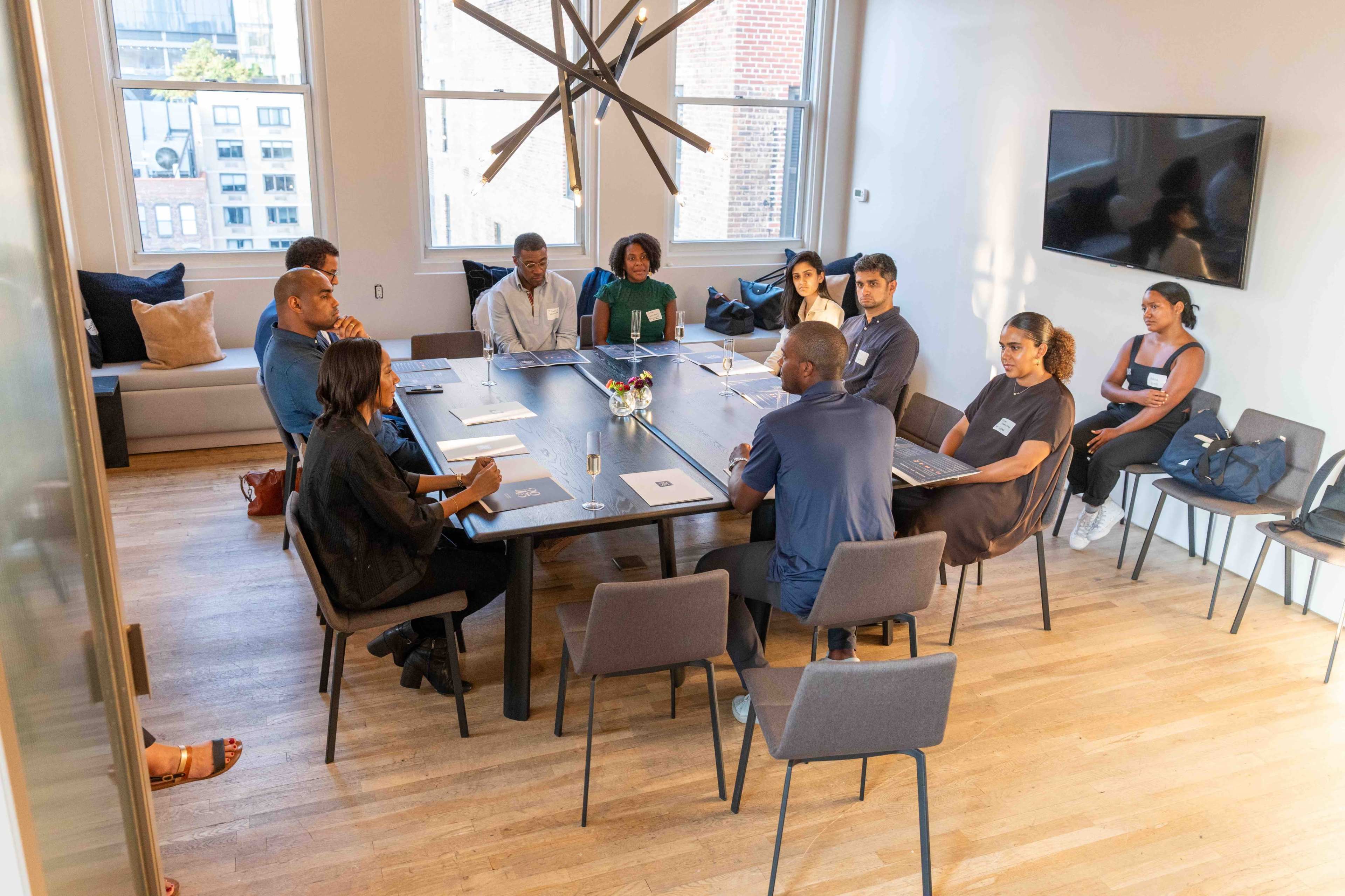 A group of twelve people sit around a large rectangular table in a well-lit meeting room.