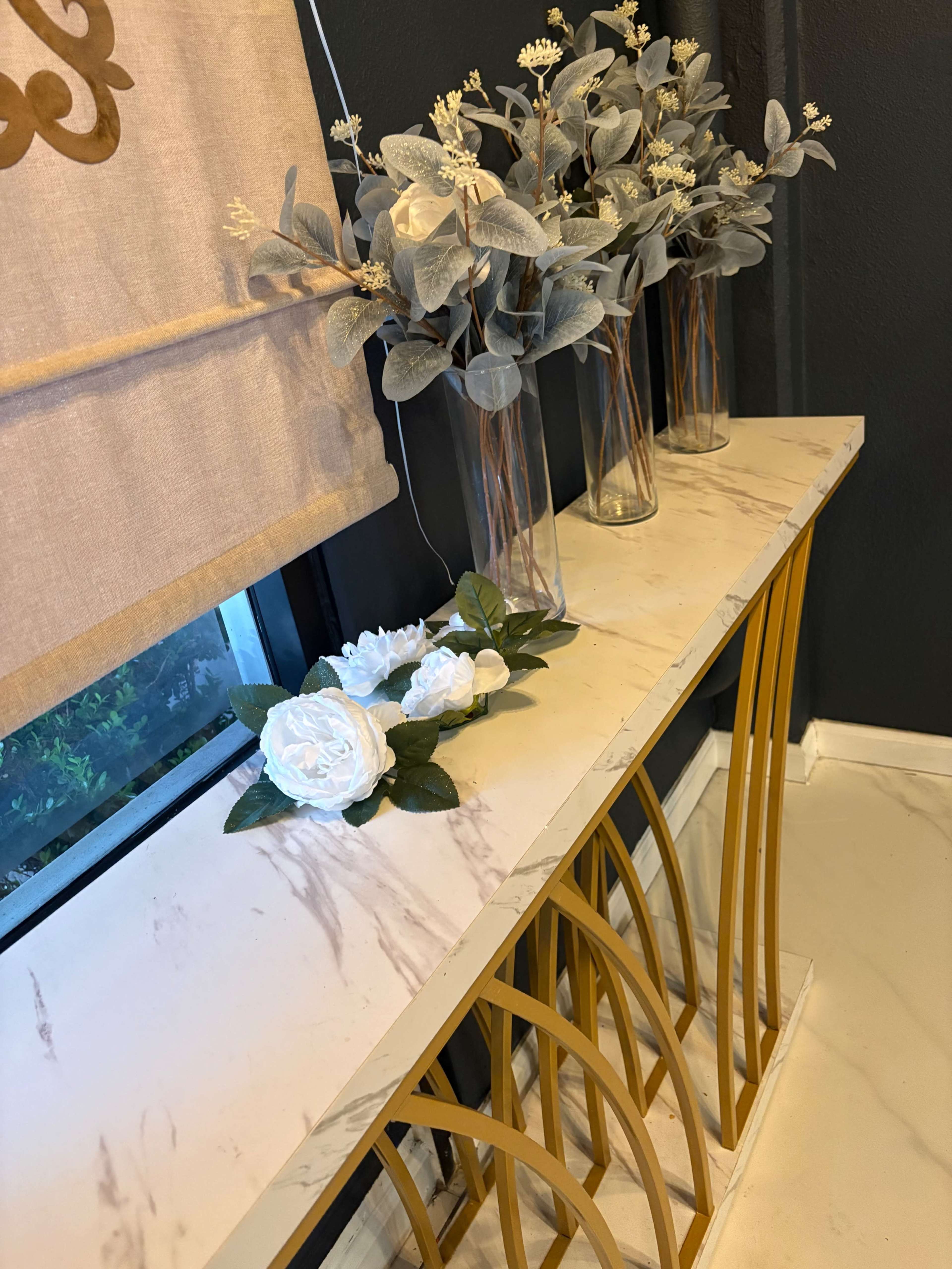 A marble-topped console table with a golden base holds two vases of faux flowers and a decorative arrangement of white floral accents.