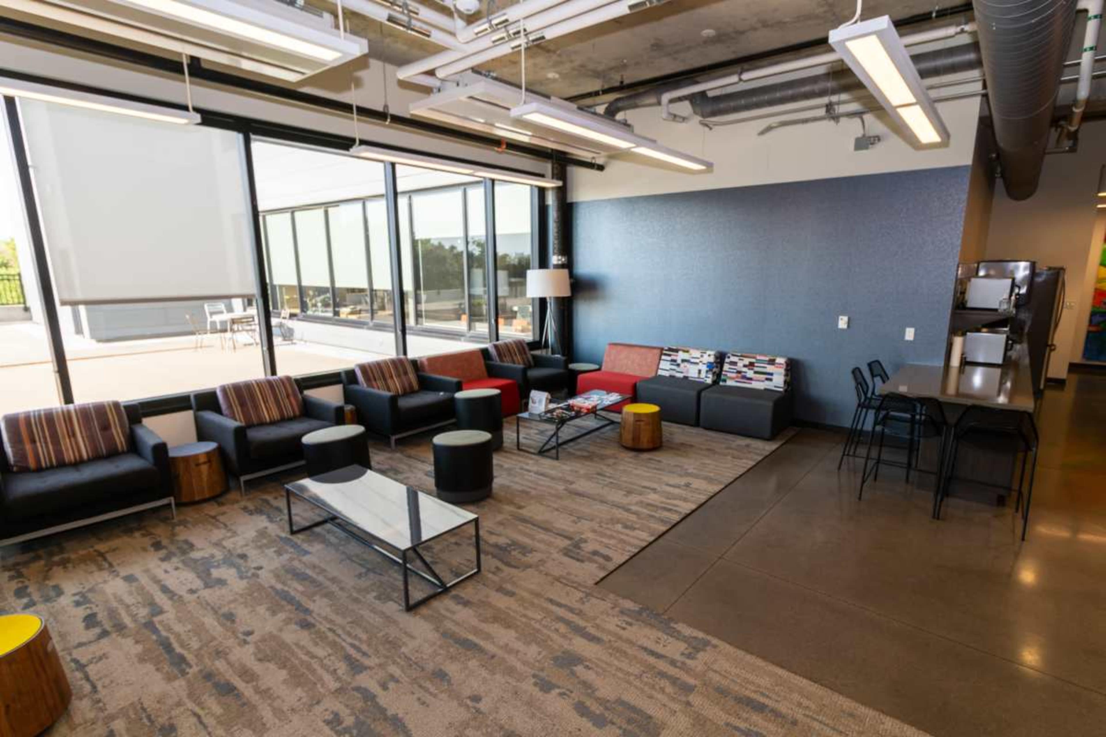 The image shows a modern lounge area featuring black sofas, a glass coffee table, and a kitchenette with barstools.