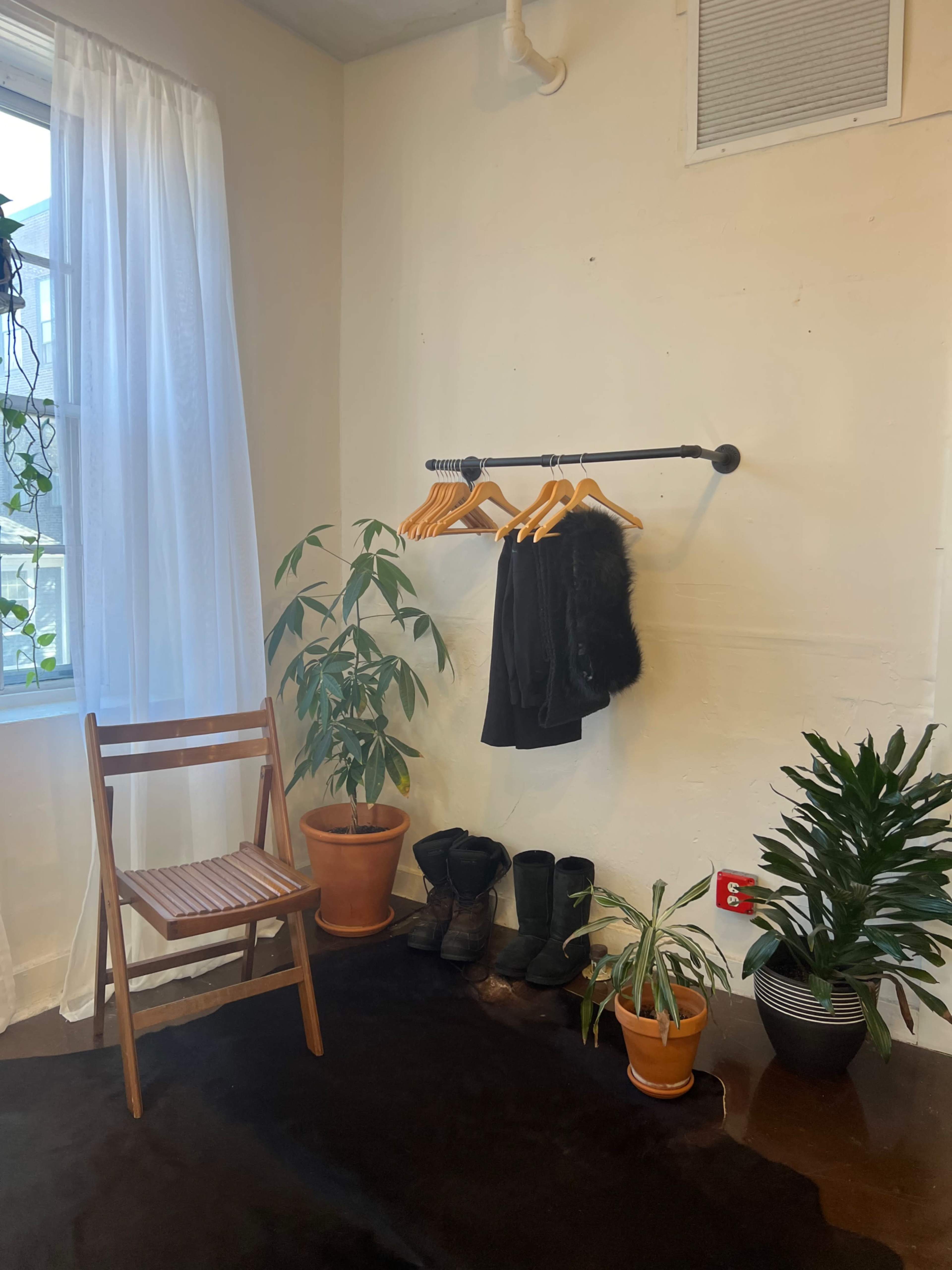 A minimalistic interior space features a chair, a clothing rack with several hangers, and various potted plants beside a window.