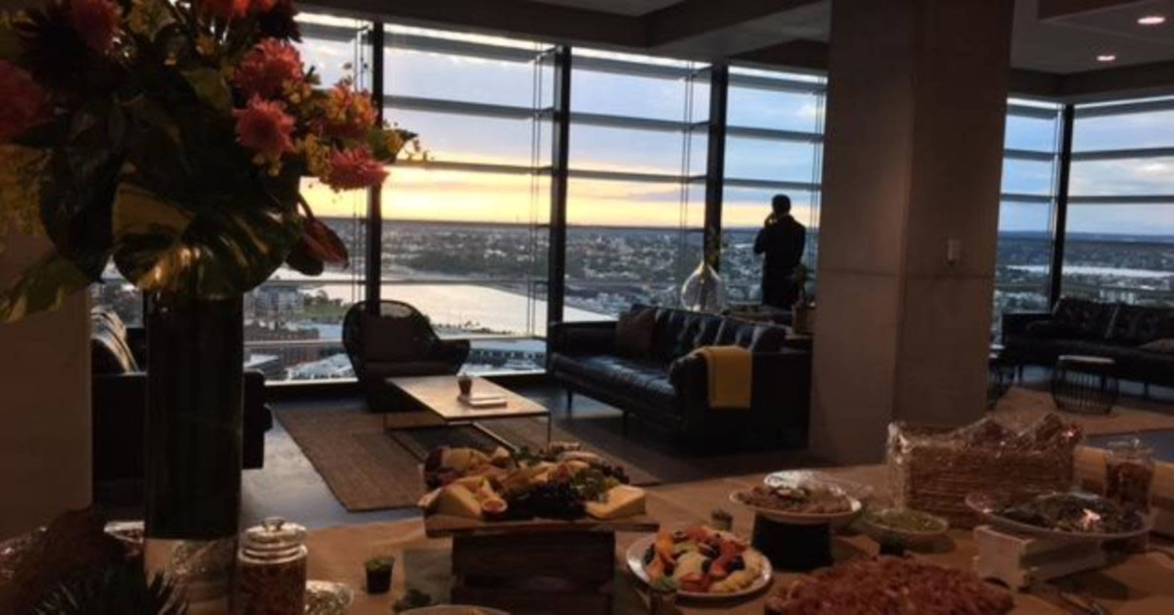 A modern interior with a buffet spread in the foreground, large windows displaying a sunset view over a body of water, and a figure standing by the window.