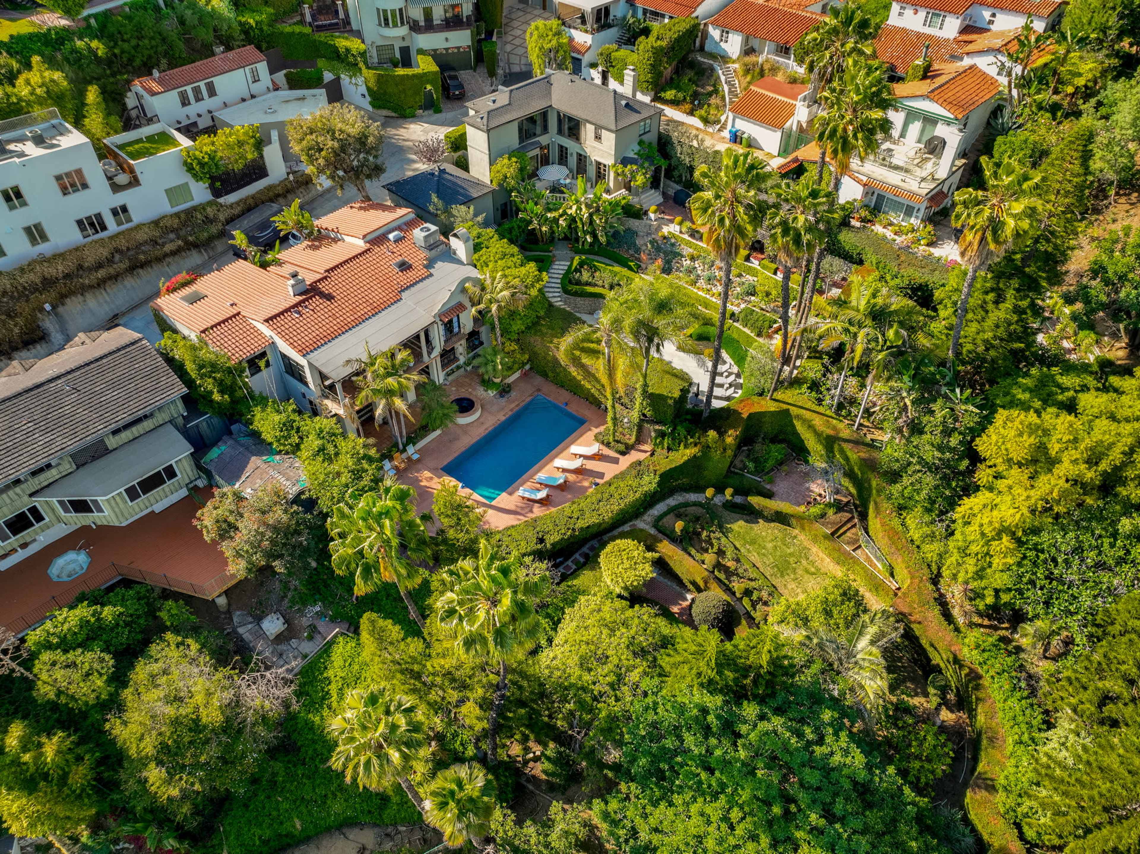 An aerial view of a residential area featuring a house with a pool surrounded by landscaped gardens and palm trees.