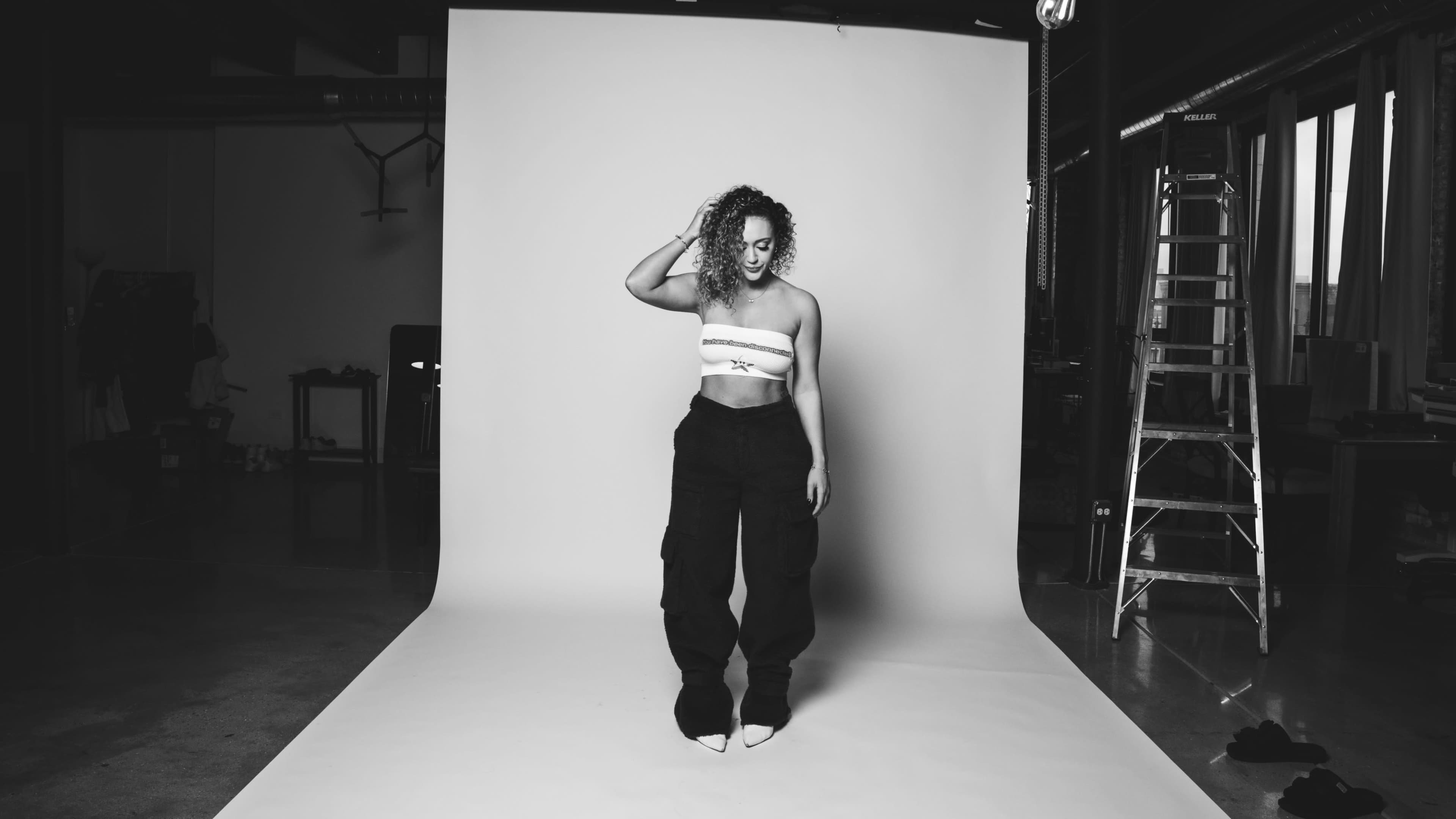 A person poses in a studio with a plain backdrop, wearing a strapless top and wide-leg pants.