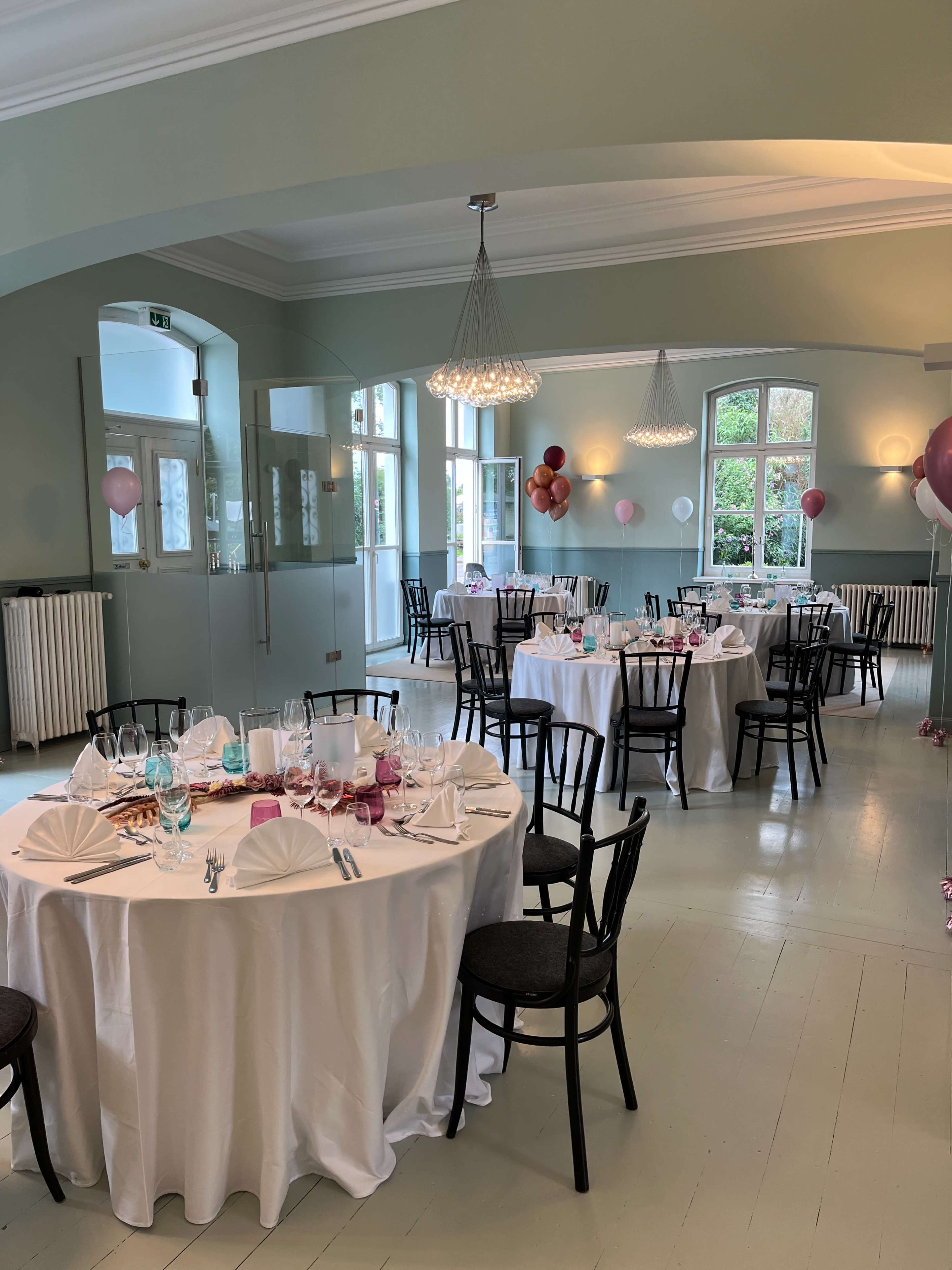 A banquet hall set for a dinner with round tables covered in white tablecloths and decorated with folded napkins, surrounded by balloons hung from the walls.