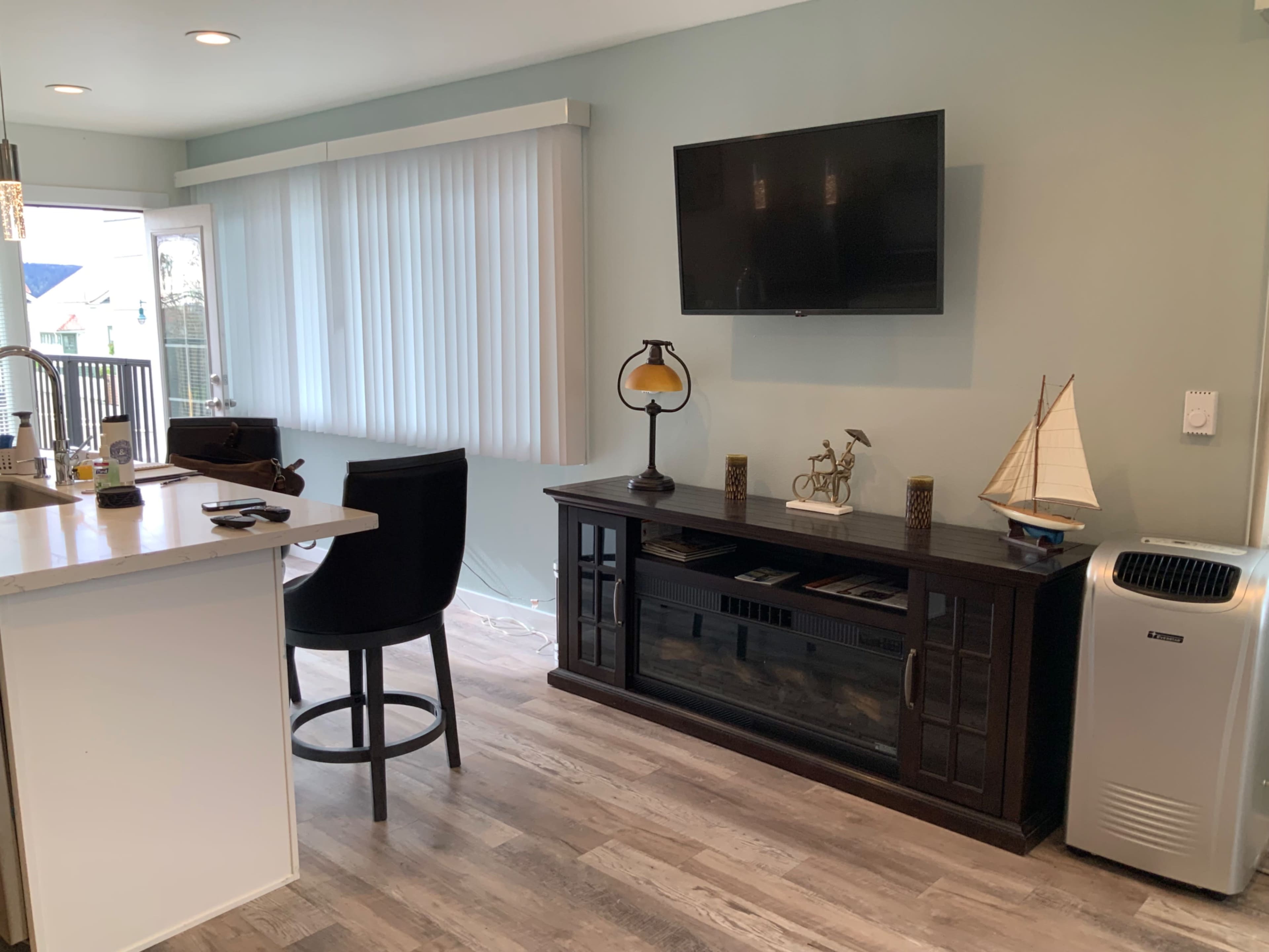 A modern living space with a television mounted on the wall, a dark wooden entertainment console, a dining area with a high-backed chair, and a window with vertical blinds.