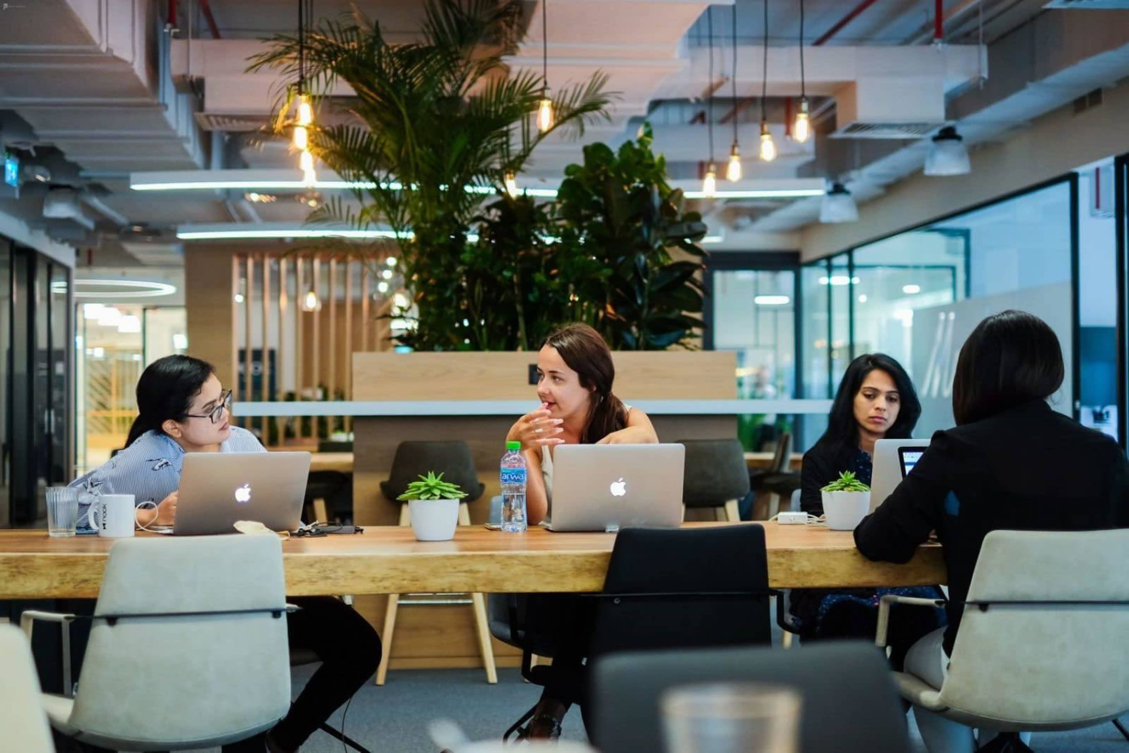 Four women sit around a wooden table in a modern office space, each engaged with their laptops and discussing ideas.
