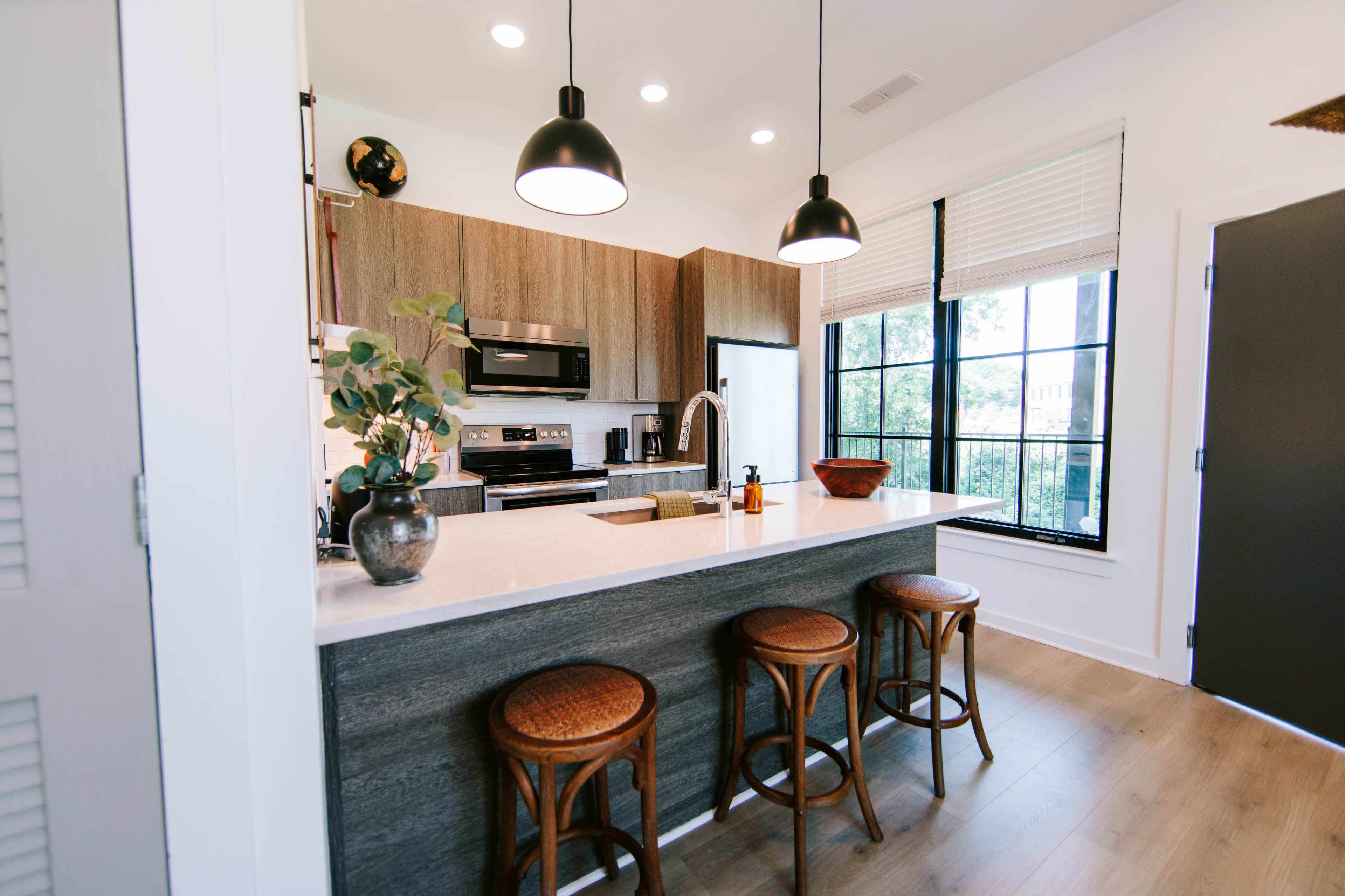 The image shows a modern kitchen with wooden cabinetry, a marble countertop, three wicker stools, and large windows letting in natural light.