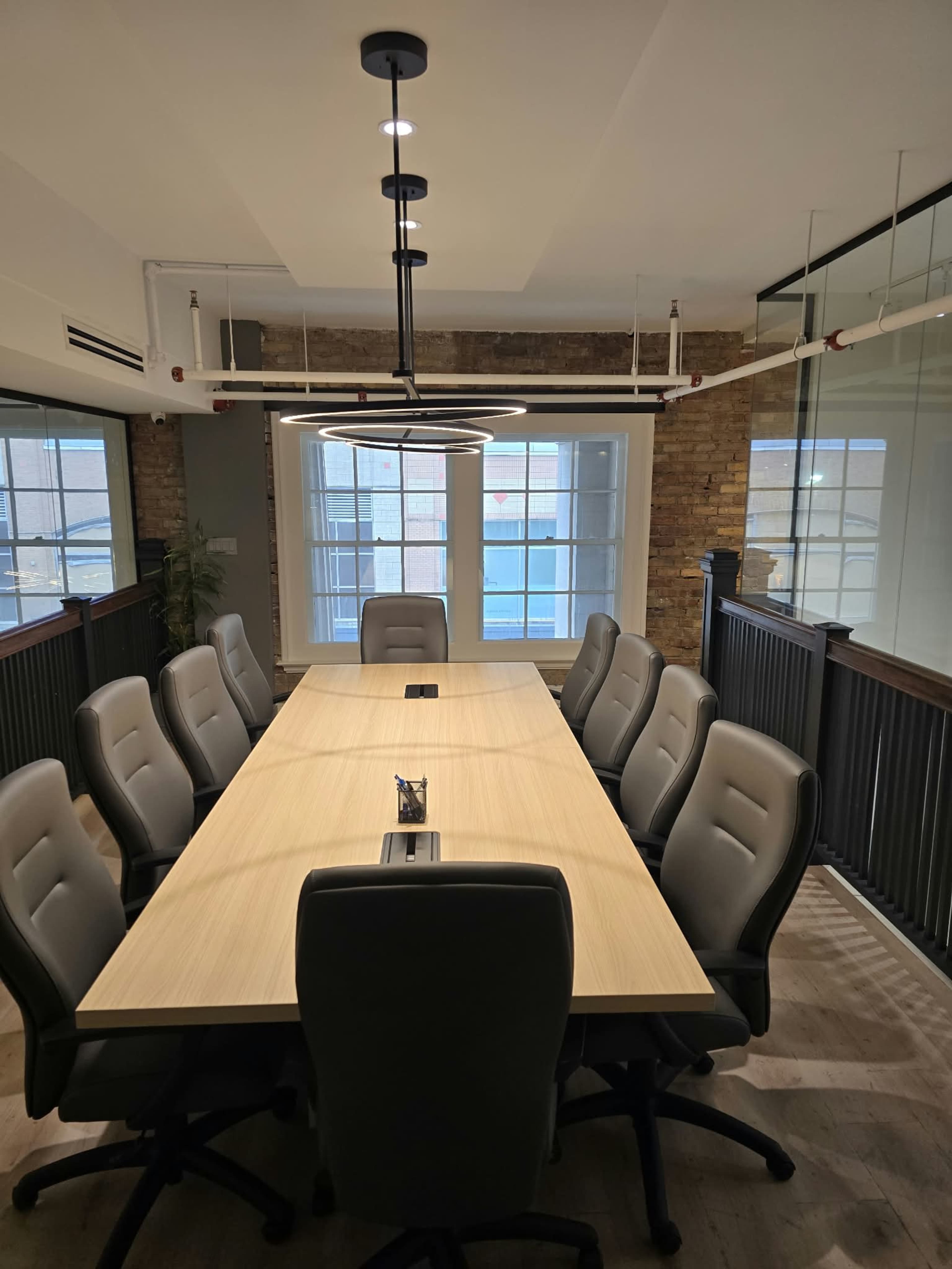 A long, rectangular conference table is surrounded by office chairs in a well-lit meeting room featuring large windows and a modern design.