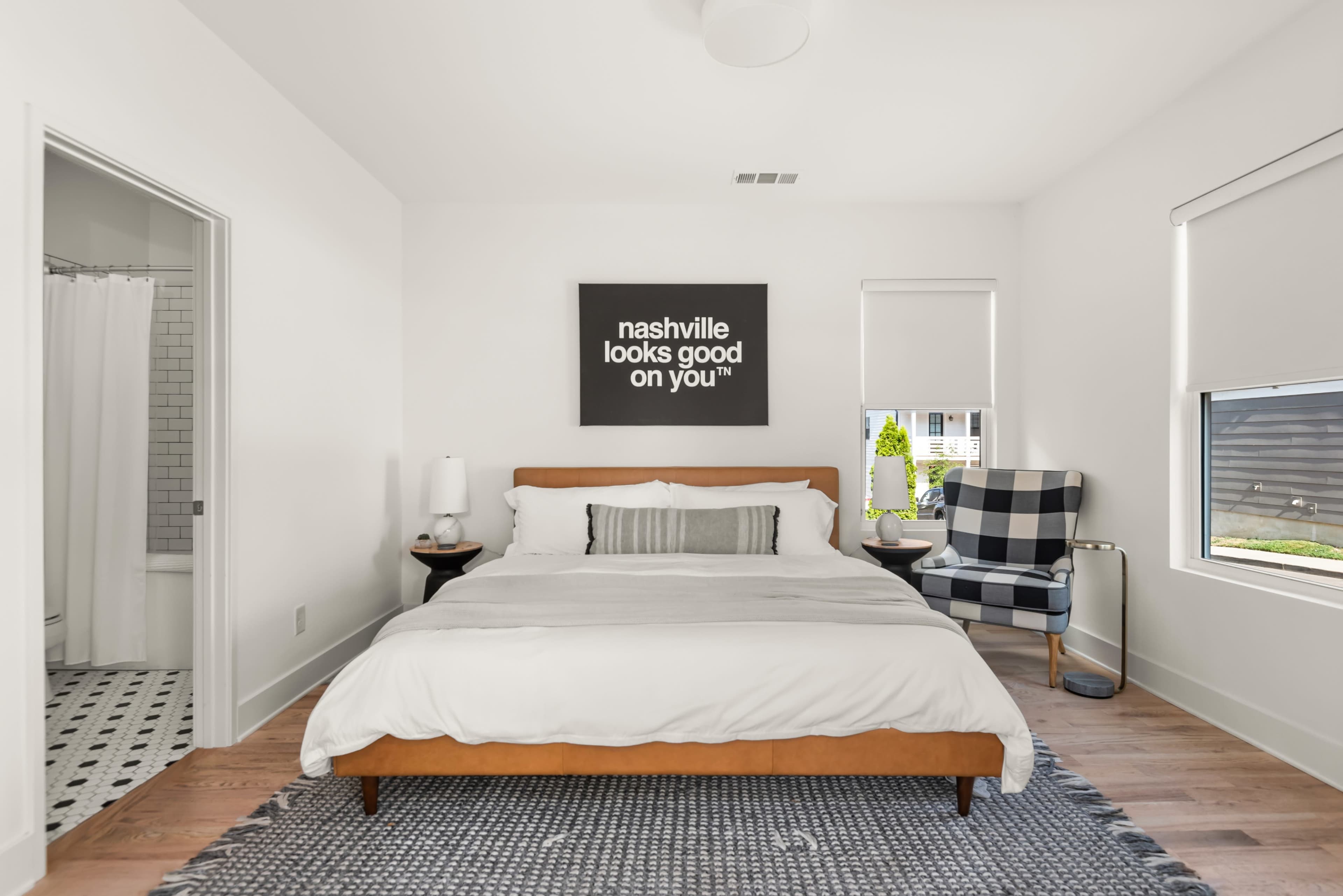 A modern bedroom features a wooden bed with a white comforter, a black-and-white checkered chair, and a wall art piece that reads "nashville looks good on you."