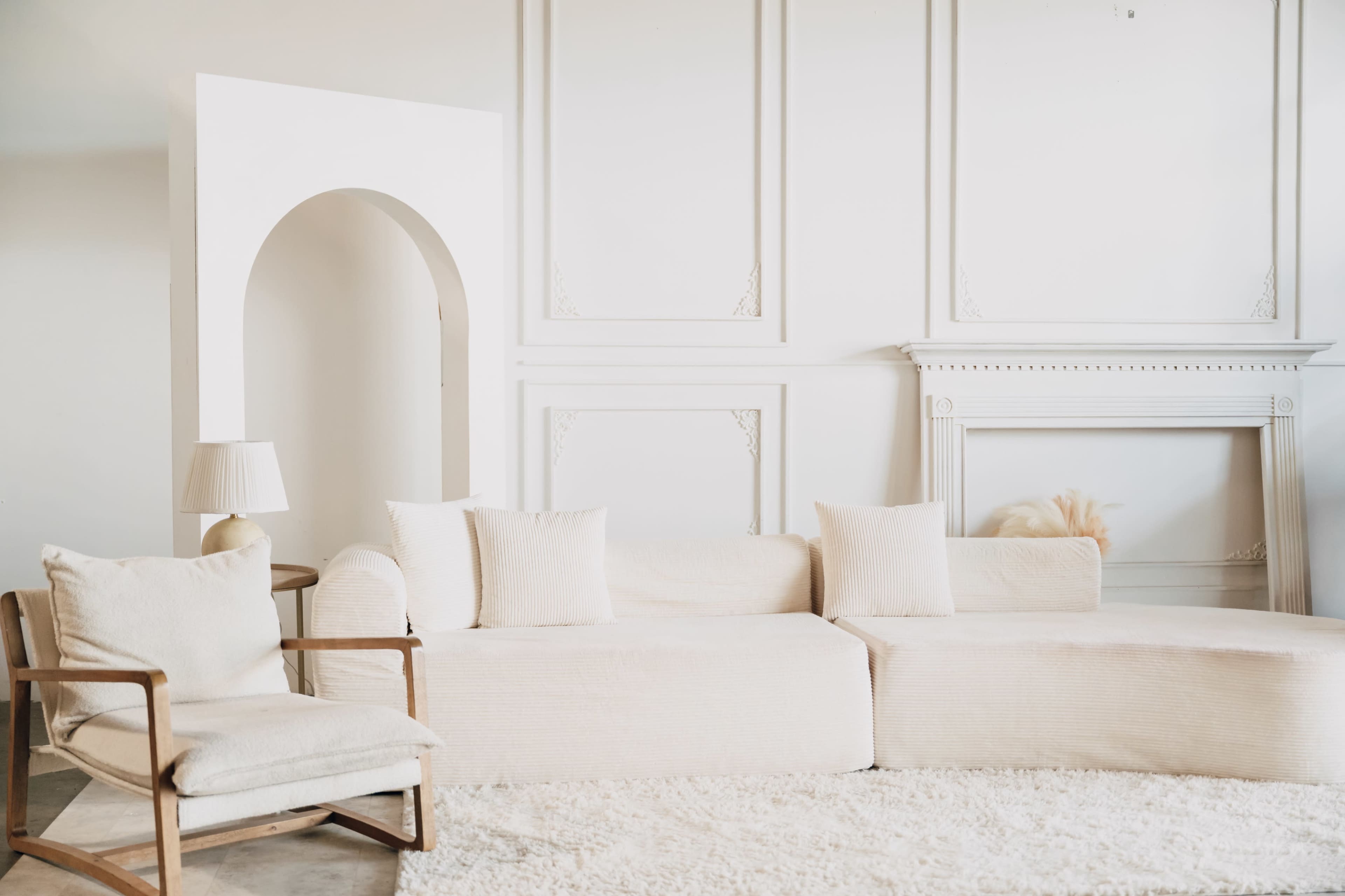The image shows a minimalist living room featuring a large beige sectional sofa, a light armchair, and a white rug, with an archway and a decorative fireplace in the background.